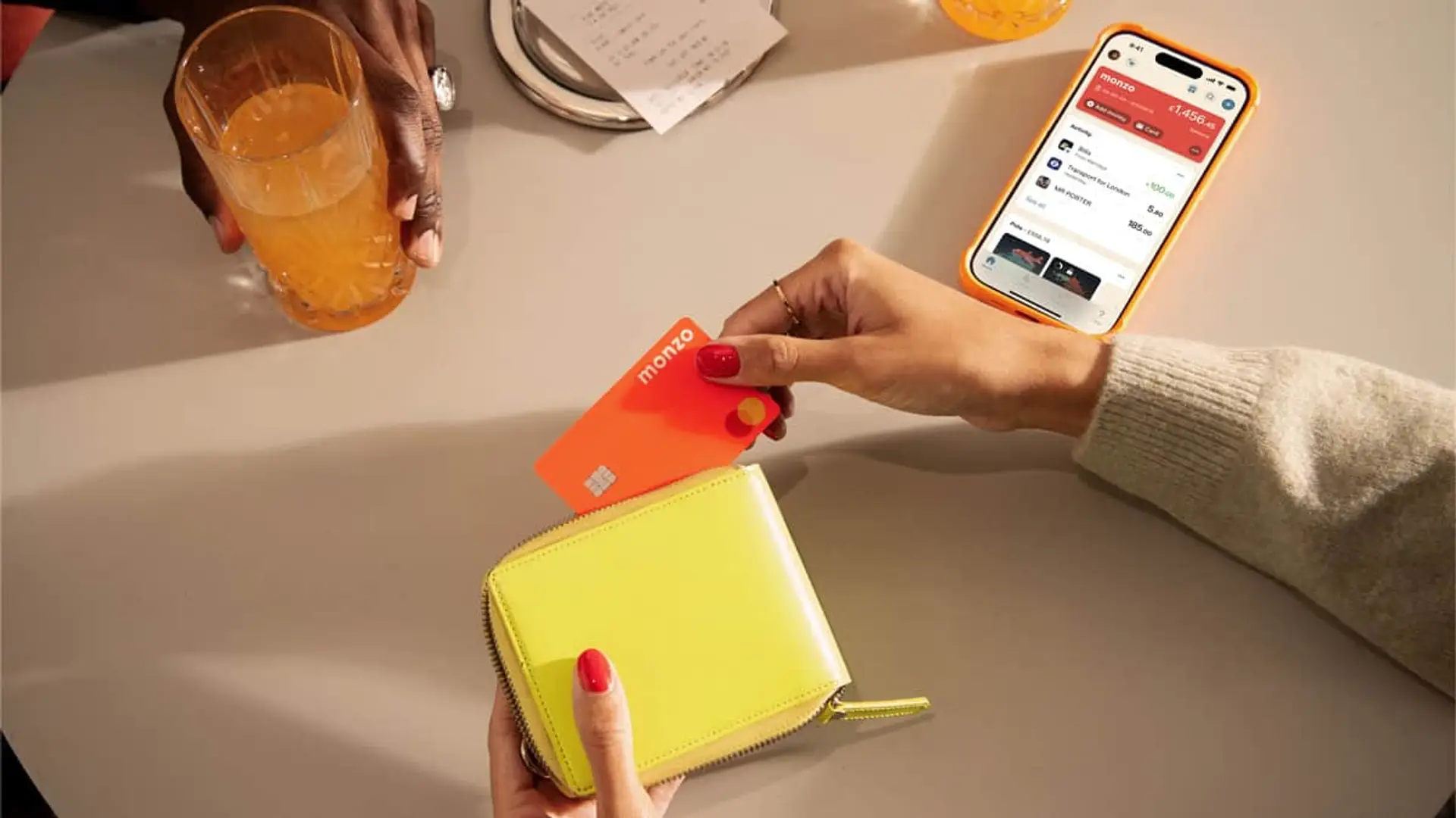 Mano sacando una tarjeta Monzo de un monedero junto a un móvil con la app abierta de banca digital.