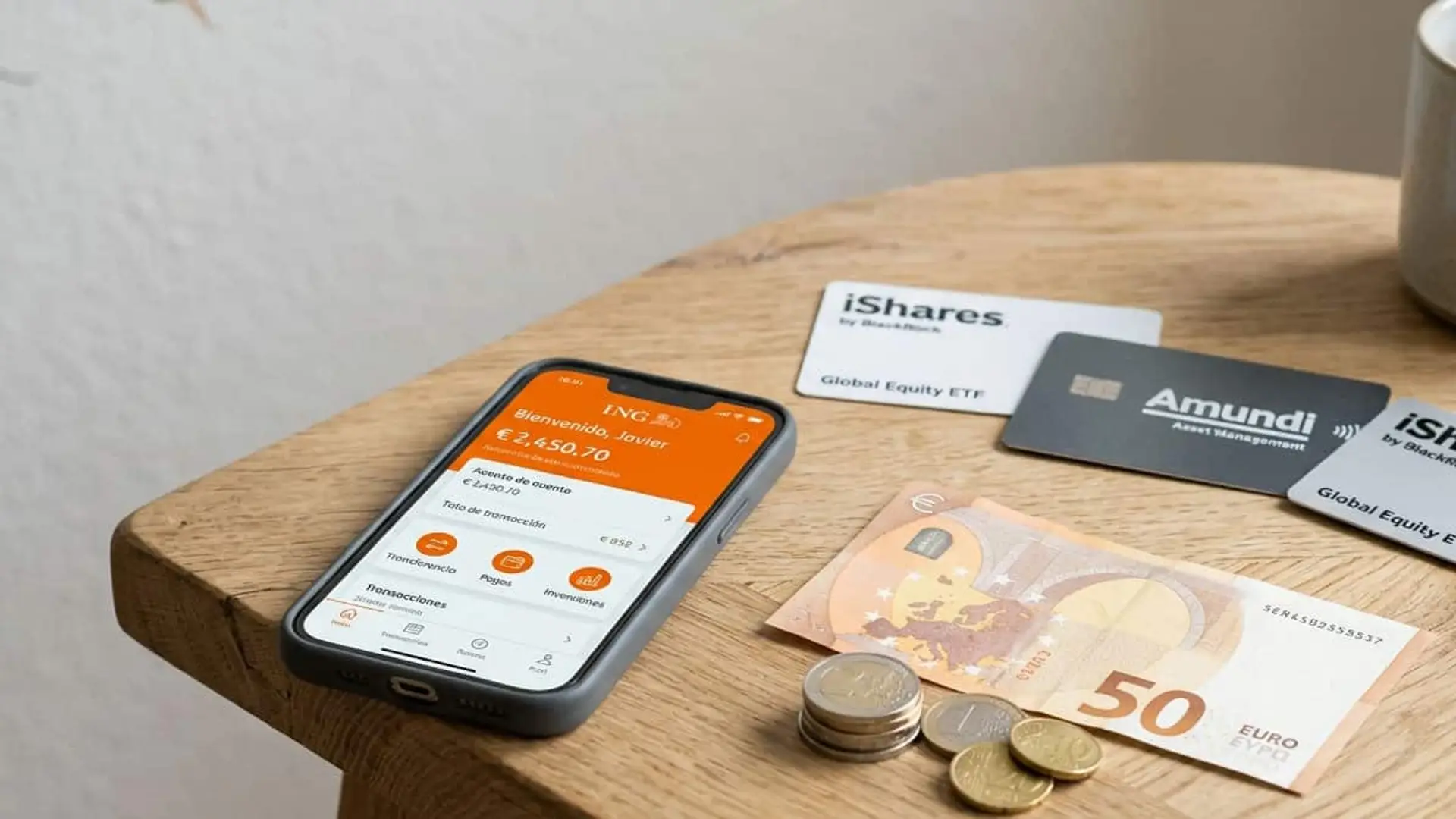 Móvil con la aplicación de ING junto a tarjetas de ETF de iShares y Amundi, billetes de 50 euros y monedas sobre una mesa.