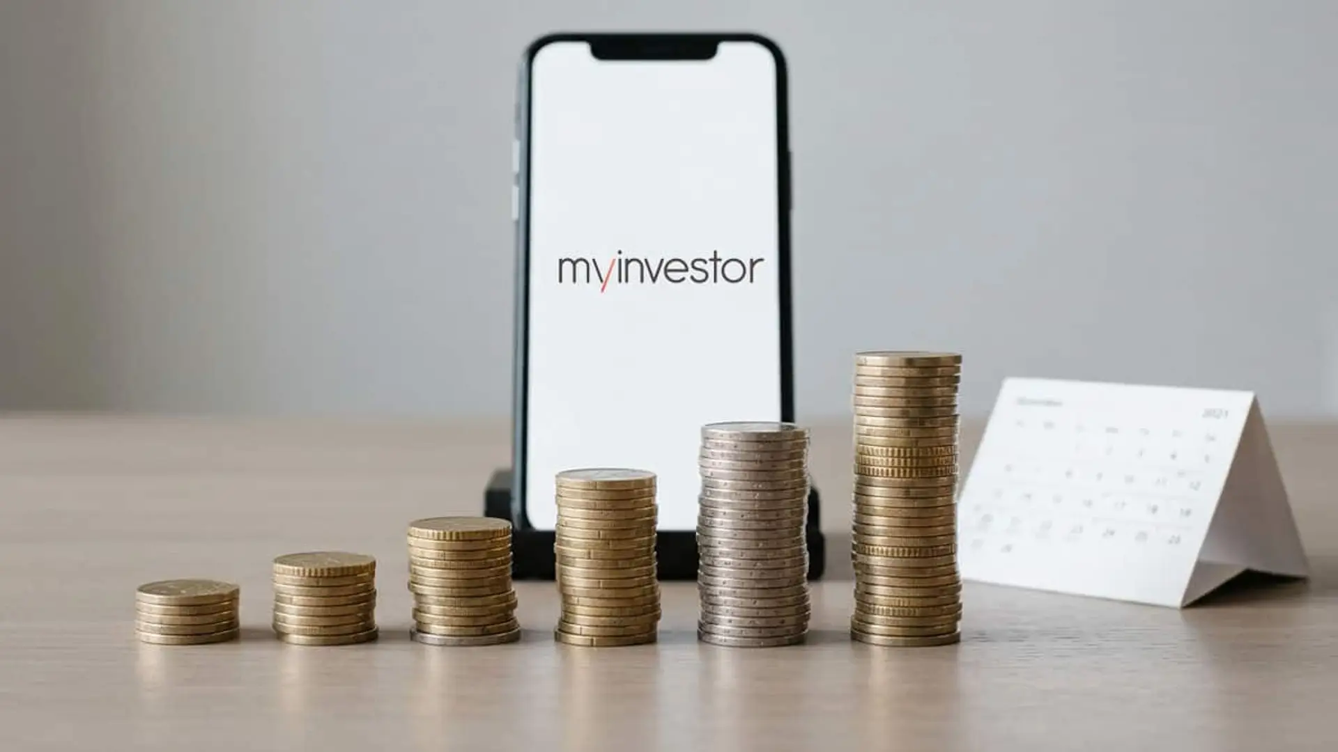 Teléfono móvil con el logotipo de MyInvestor junto a seis pilas de monedas de altura creciente y un calendario de sobremesa.