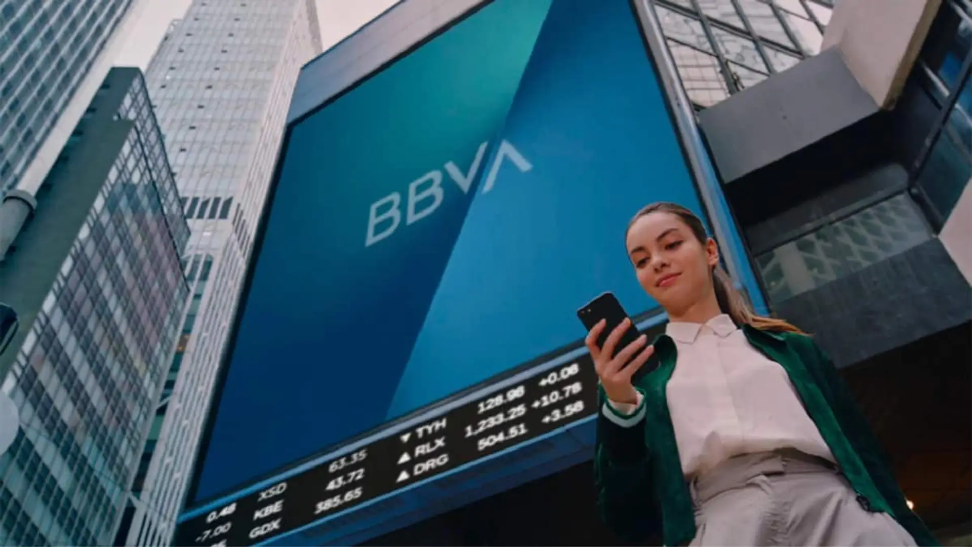 Mujer mirando el móvil frente a una gran pantalla con el logotipo de BBVA en un entorno urbano