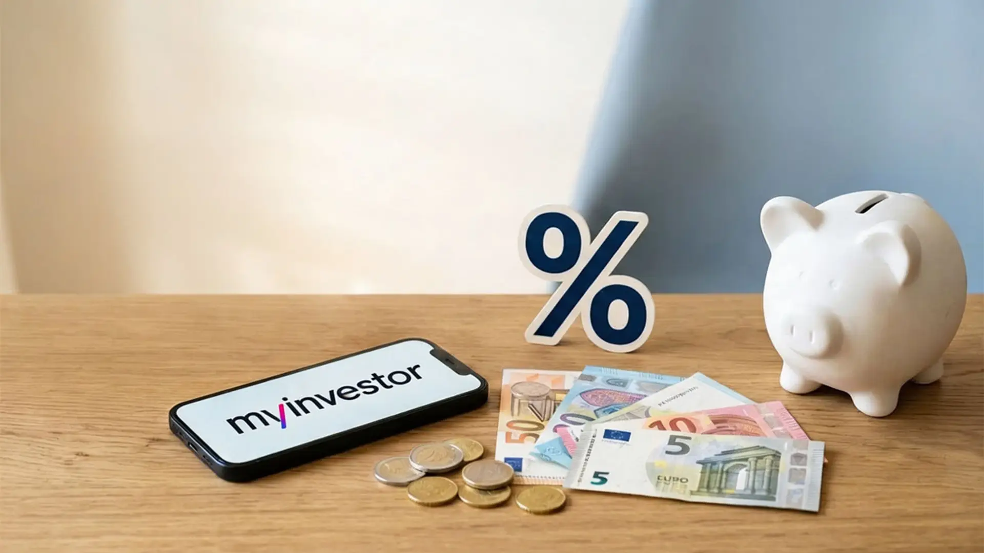 Teléfono móvil con el logo de MyInvestor junto a una hucha blanca, billetes, monedas y un símbolo de porcentaje sobre una mesa