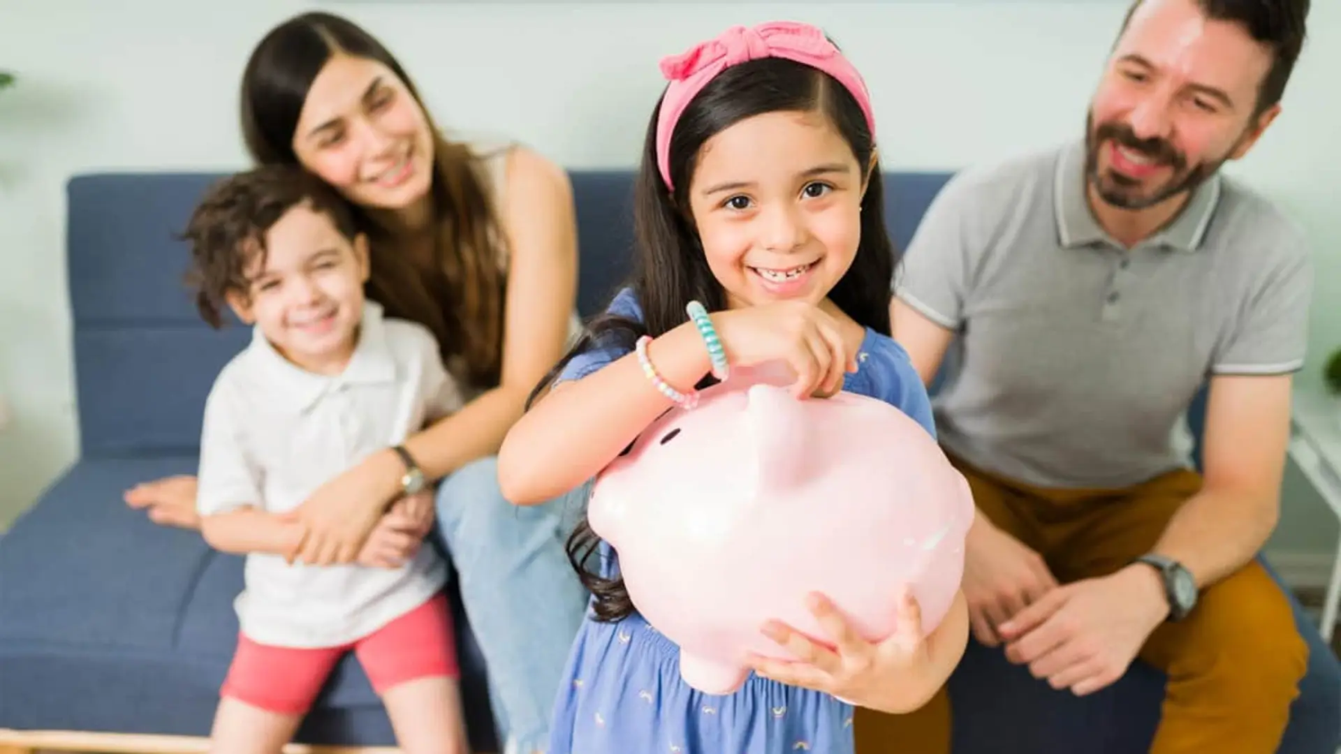 España se anima a invertir: el patrimonio financiero de las familias crece más que en el resto de Europa Niña sonriendo mientras introduce una moneda en una hucha con forma de cerdito, junto a su familia en el sofá.