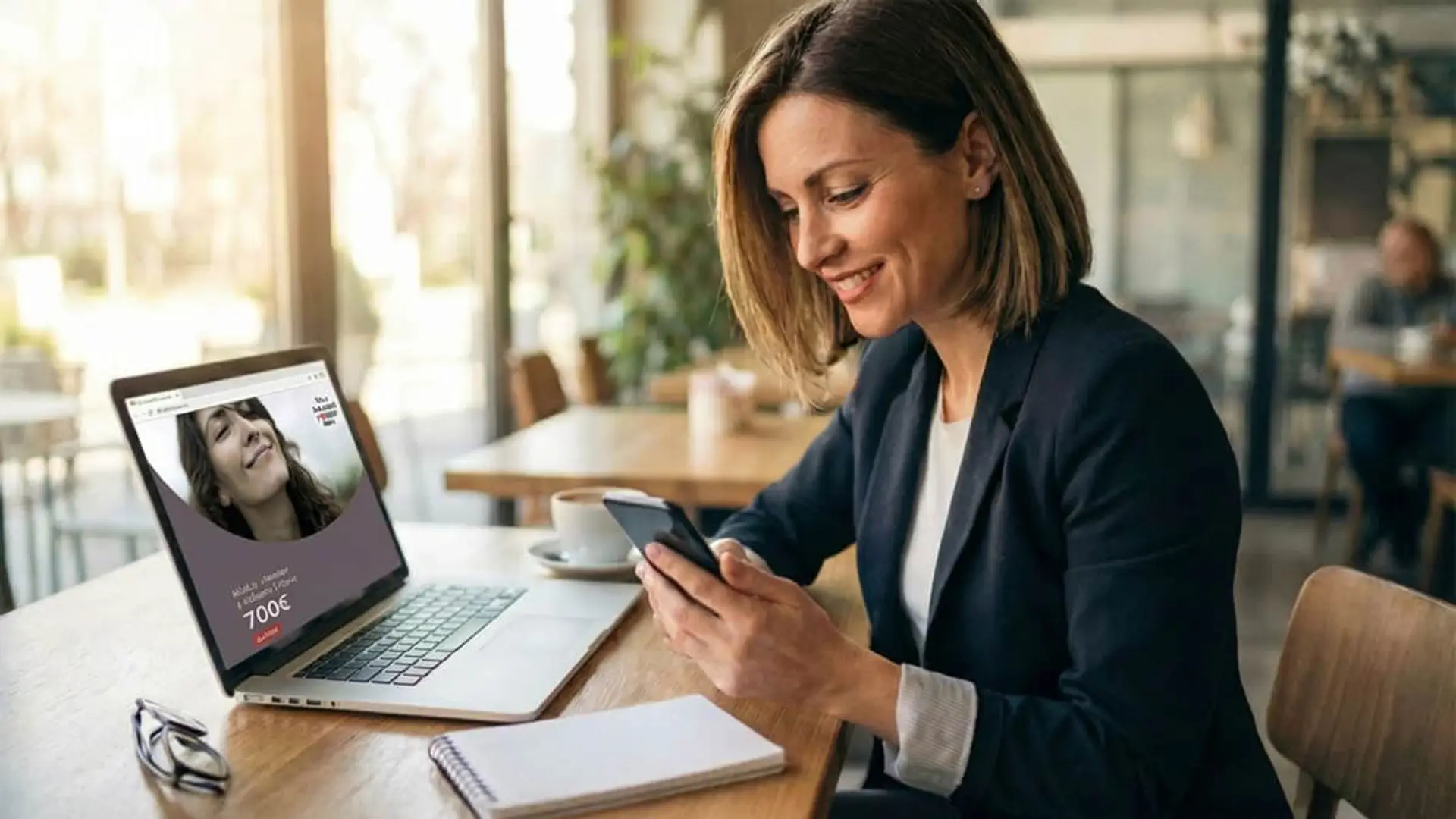 Mujer consultando oferta de 700€ de Kutxabank en el móvil y portátil en mesa de cafetería.