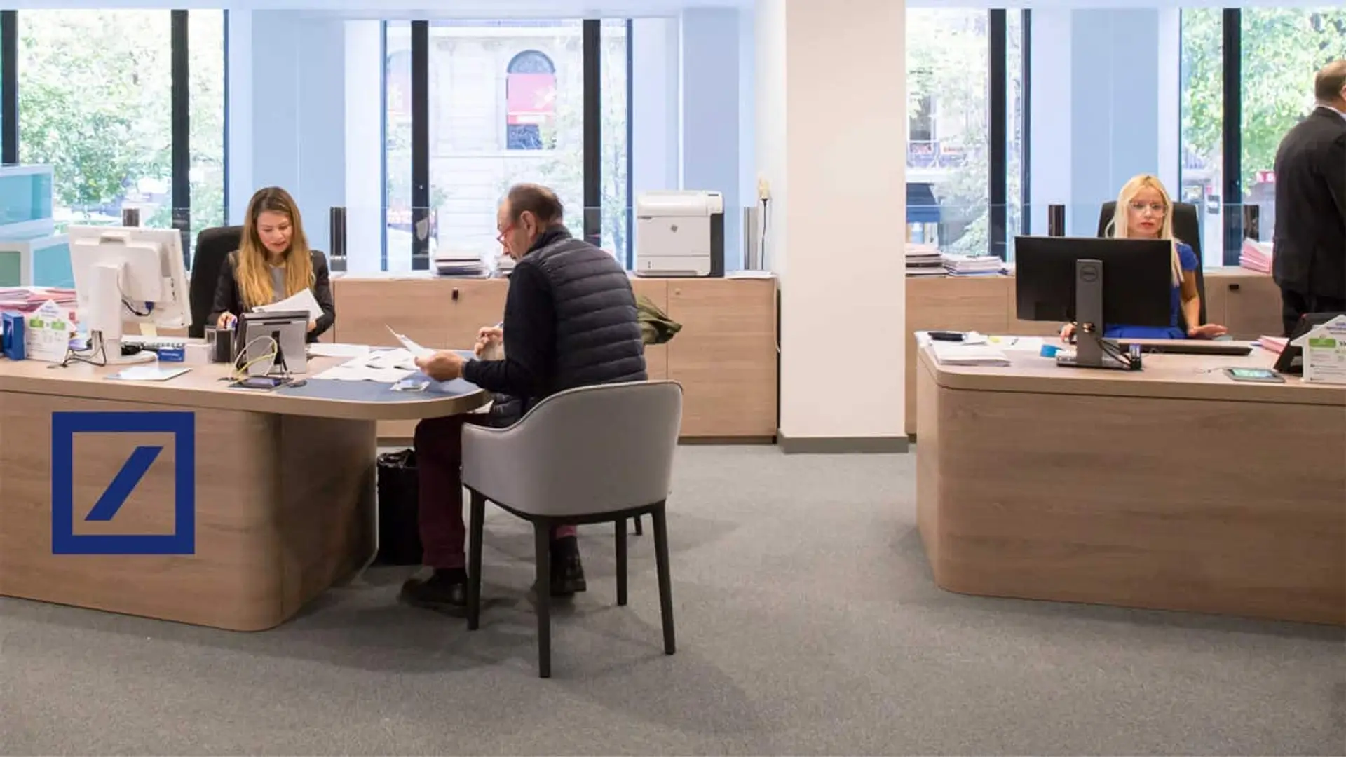 Interior de una oficina de Deutsche Bank, con empleados atendiendo a clientes en mesas modernas y grandes ventanales al fondo.