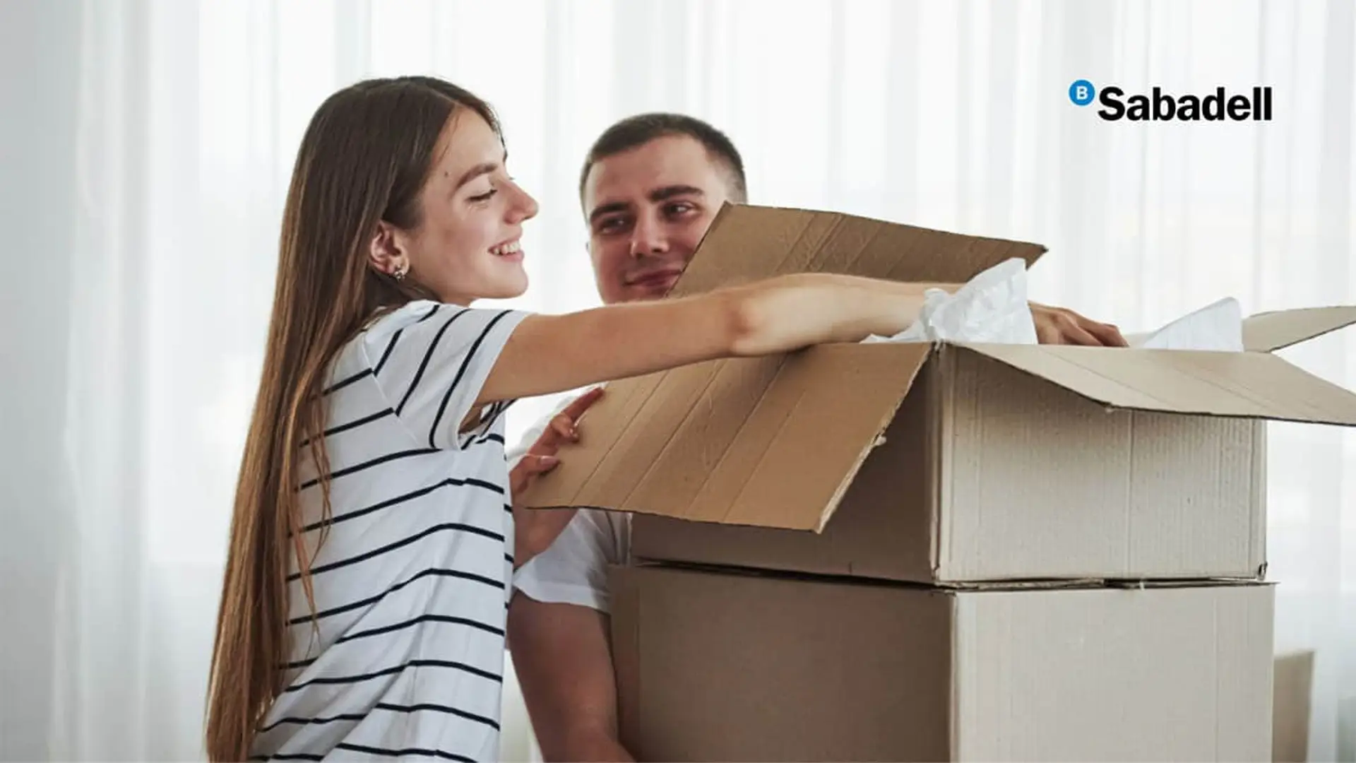 Banco Sabadell mueve ficha con sus hipotecas: sube la fija y abarata la variable Pareja joven feliz desempaquetando cajas de cartón en su nueva casa con logo de Banco Sabadell.