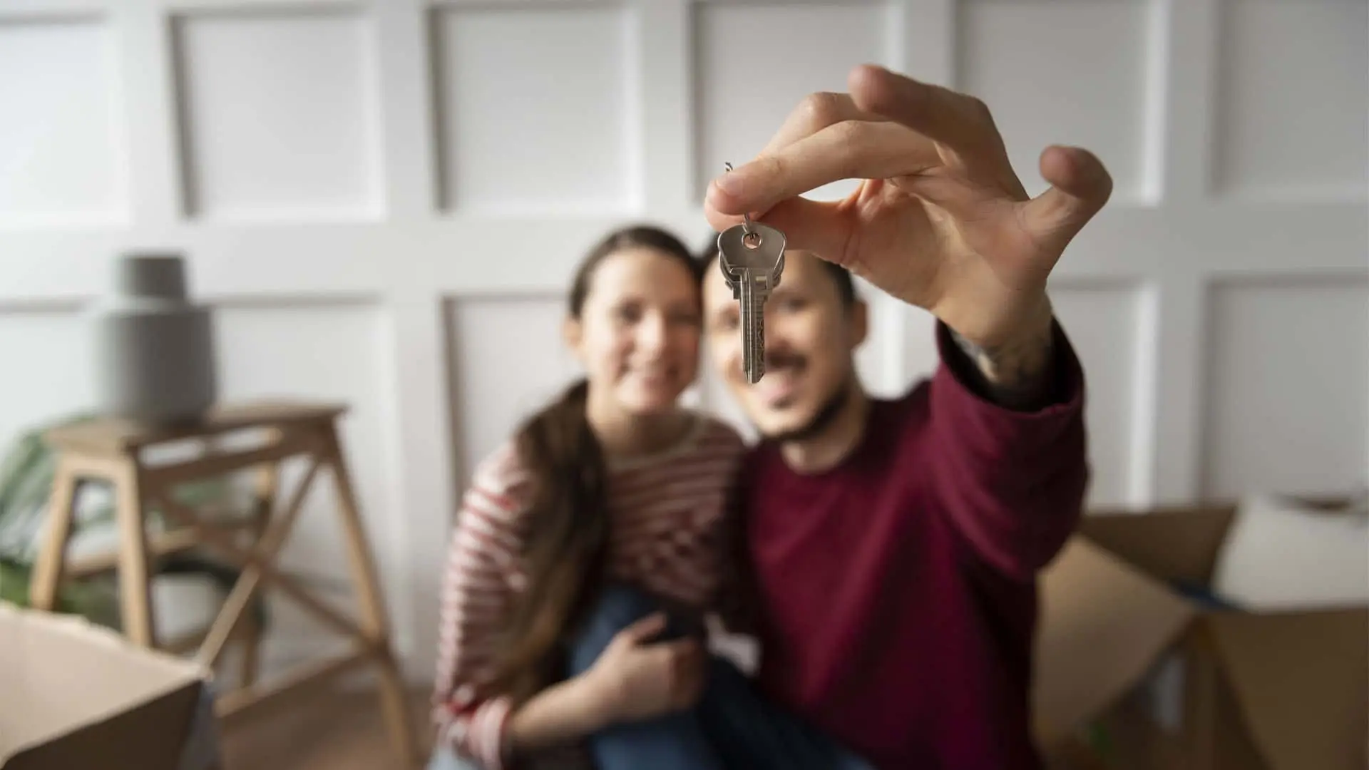 Pareja feliz mostrando las llaves de su nueva casa, con las cajas de la mudanza de fondo, tras firmar la hipoteca.