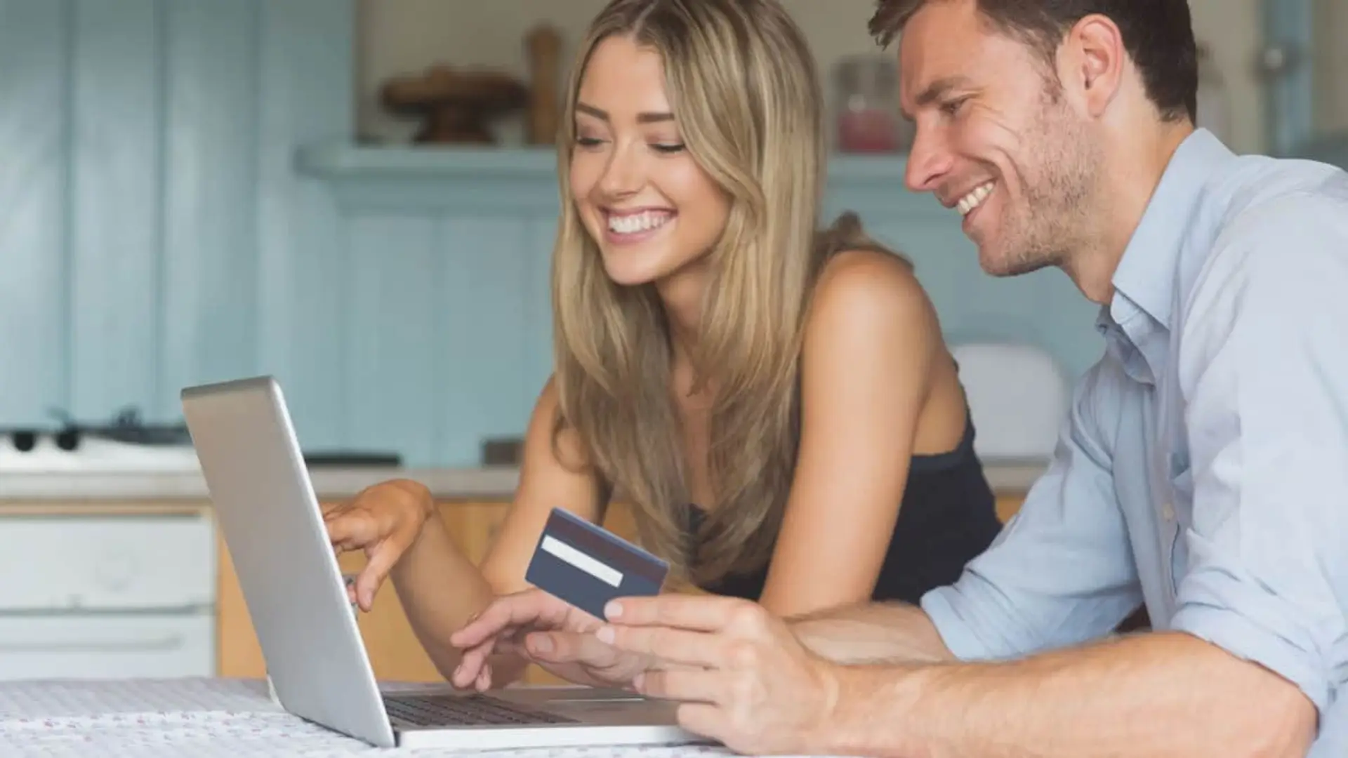 Pareja joven sonriente realizando gestión online con portátil y tarjeta bancaria.