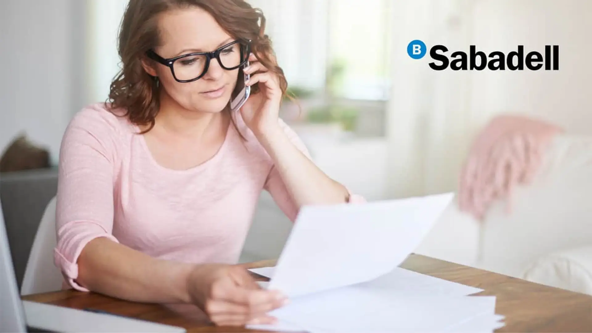 Banco Sabadell sube comisiones este verano: qué tienes que tener contratado para seguir pagando cero euros Mujer gestionando sus documentos financieros de Banco Sabadell por teléfono para resolver dudas de su cuenta.
