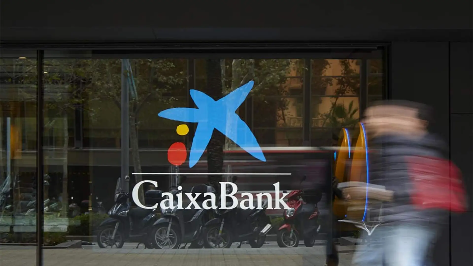 Logotipo de CaixaBank en la fachada acristalada de una oficina bancaria en una ciudad.