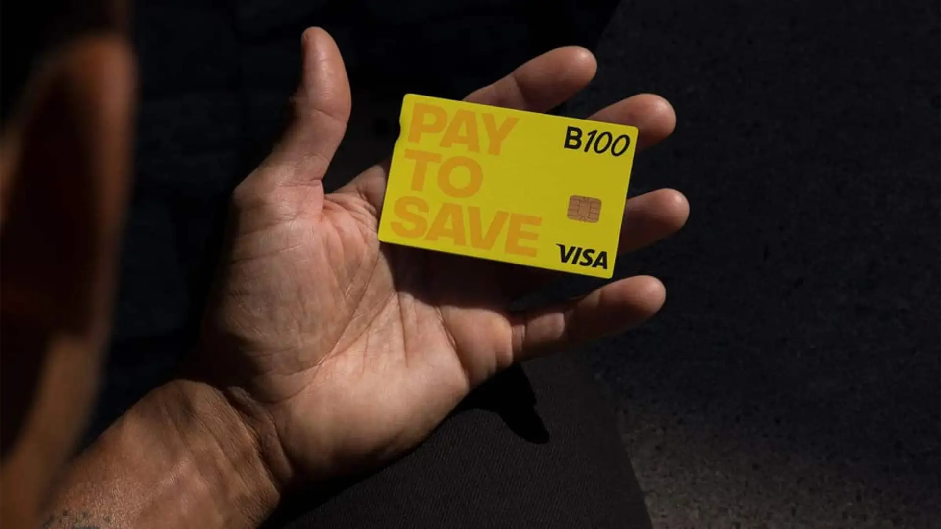 Mano sosteniendo una tarjeta bancaria B100 de Visa con el mensaje Pay to Save.