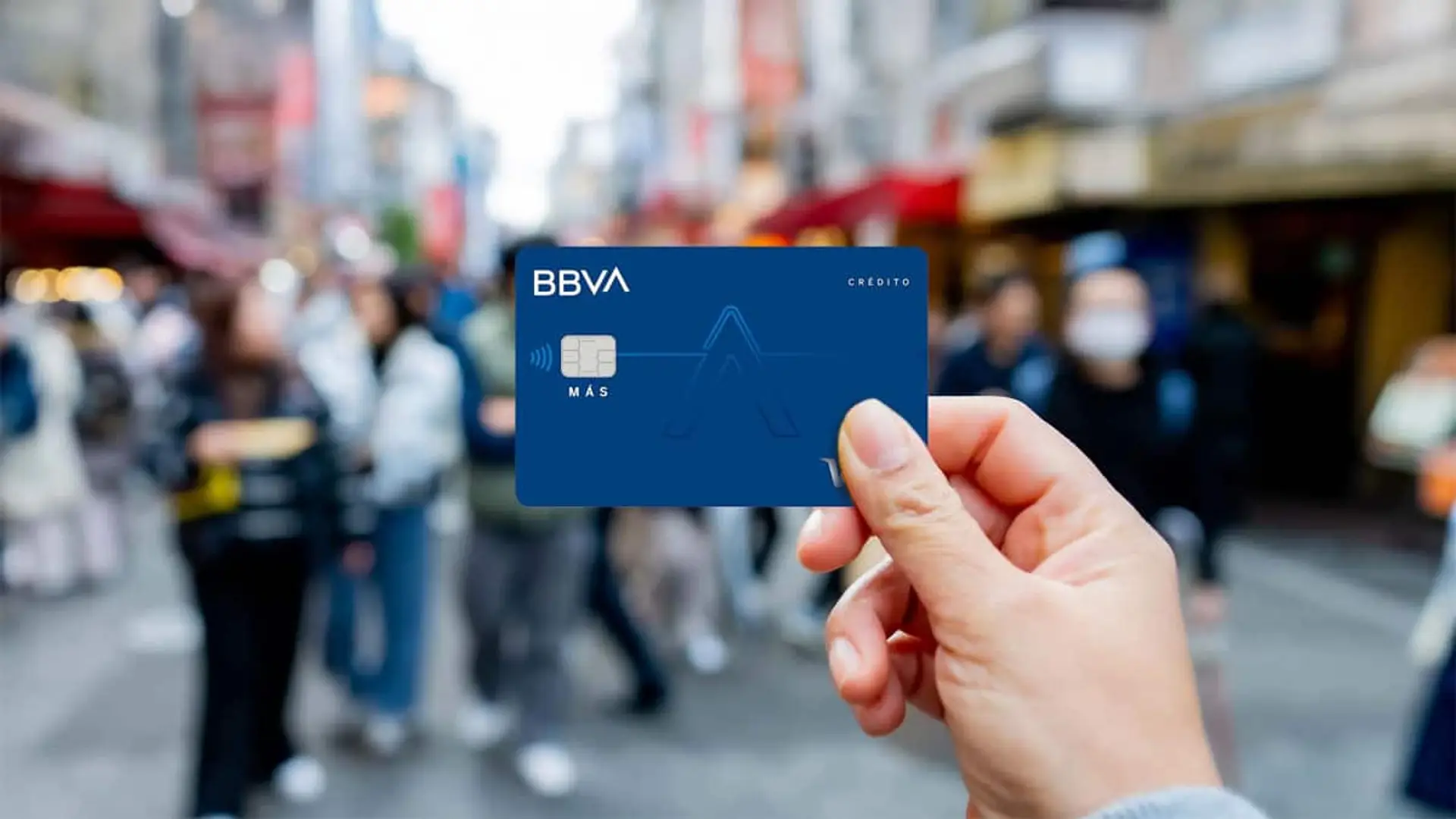 Mano sosteniendo una tarjeta de crédito BBVA Más en una calle concurrida, lista para pago contactless.