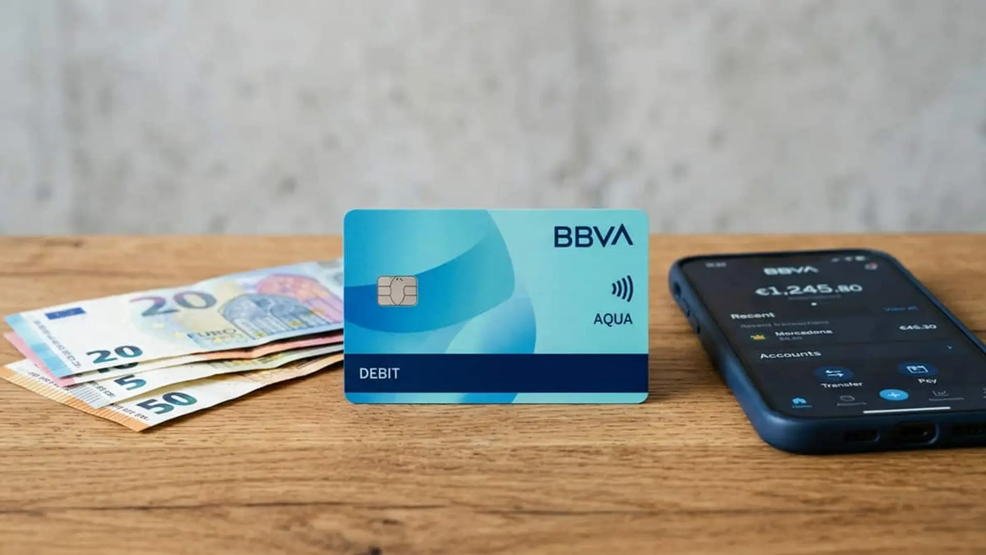 BBVA alarga hasta el 10 de noviembre su oferta de hasta 1.060 euros: los cinco bloques que la forman Tarjeta de débito Aqua de BBVA sobre una mesa de madera, junto a varios billetes de euro y un móvil con la aplicación del banco.