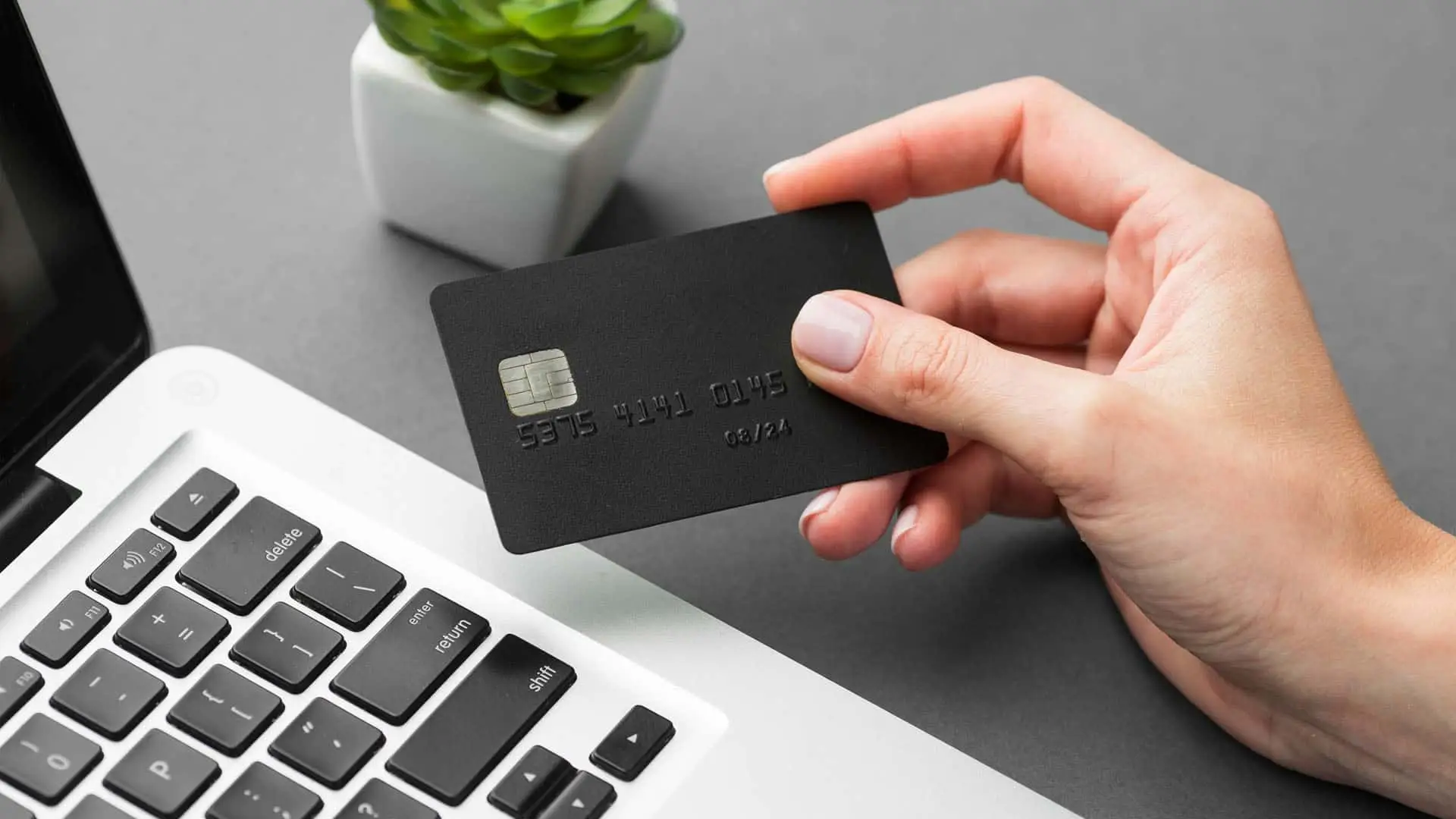 La OCU recomienda usar tarjetas de pago virtuales para evitar los fraudes online Mano sosteniendo una tarjeta bancaria negra junto a un ordenador portátil, simbolizando compras o pagos online.