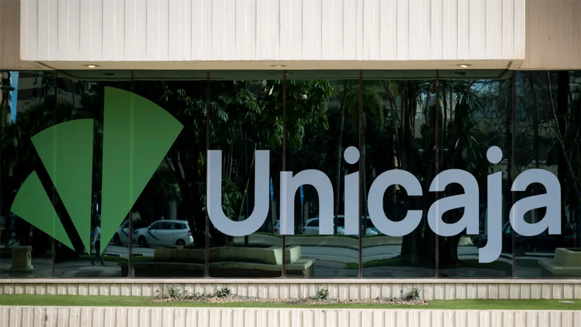 Unicaja alarga su cuenta estrella hasta el 31 de octubre Logo de Unicaja en la cristalera de una de sus oficinas o sucursales bancarias con el reflejo de la calle.