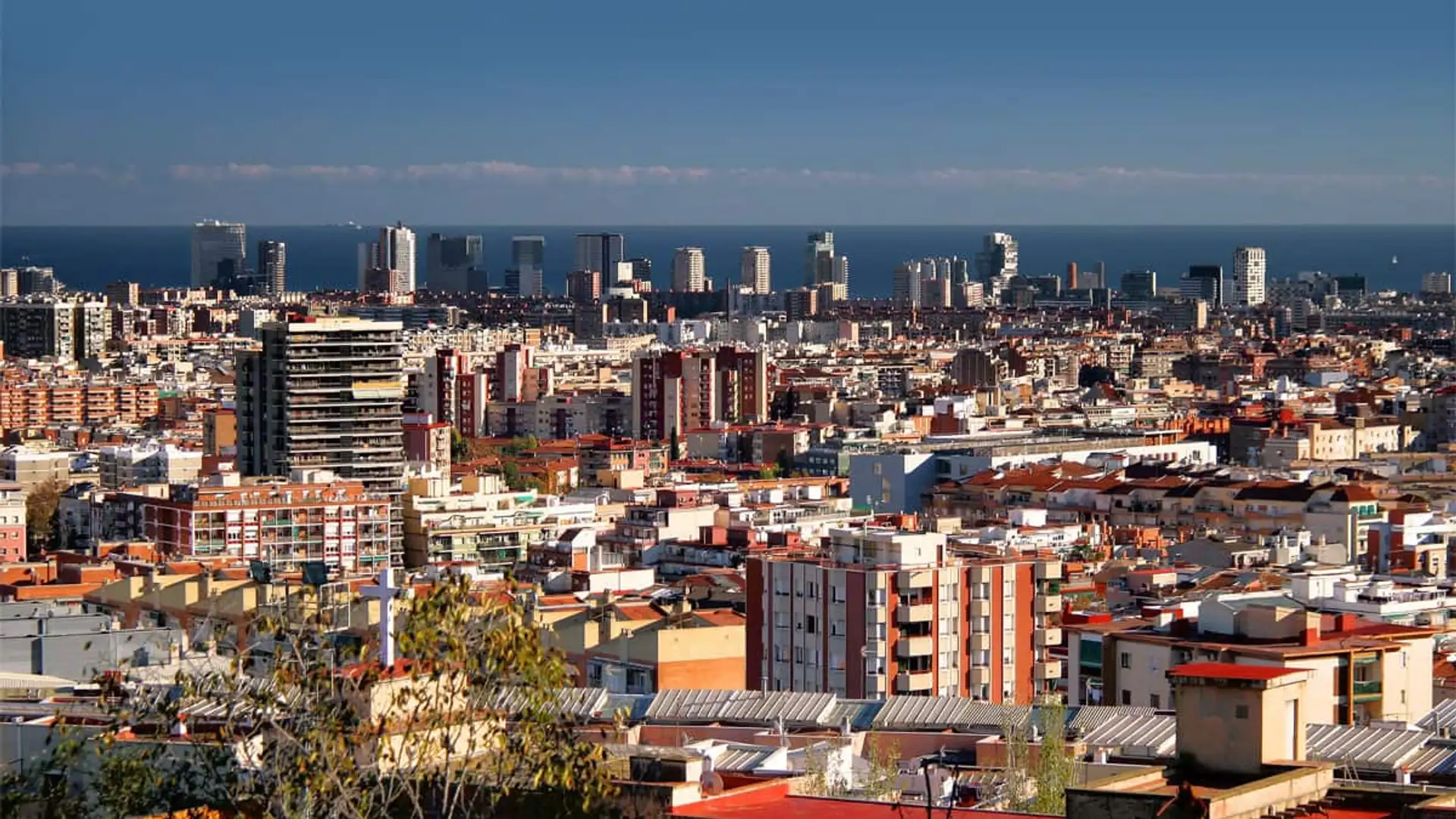 Cataluña estudia prohibir la compra de vivienda para especular: ¿adiós a los pisos de inversión? Panorámica de la ciudad de Barcelona con el mar al fondo y una multitud de bloques de pisos y viviendas.