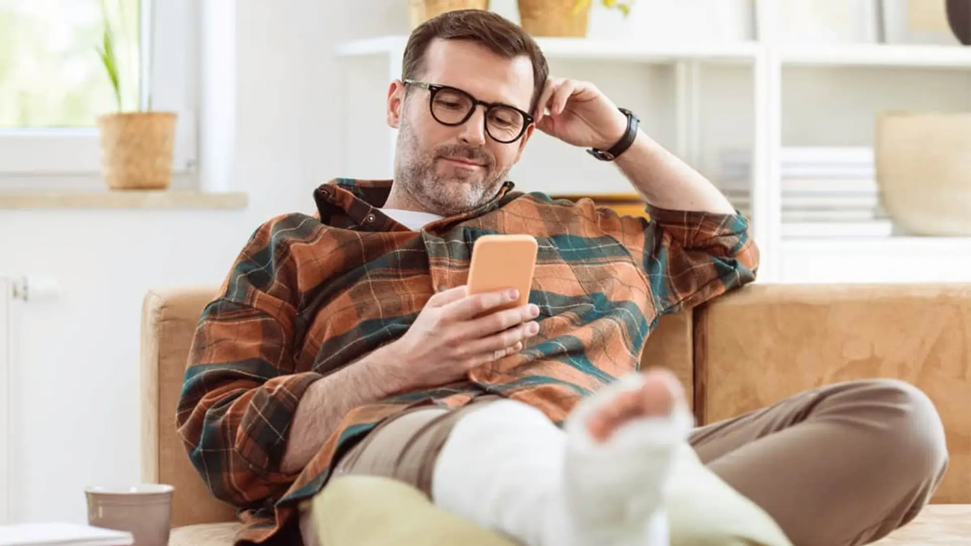 Hombre de baja con pierna escayolada gestionando trámites desde el sofá con el teléfono móvil.