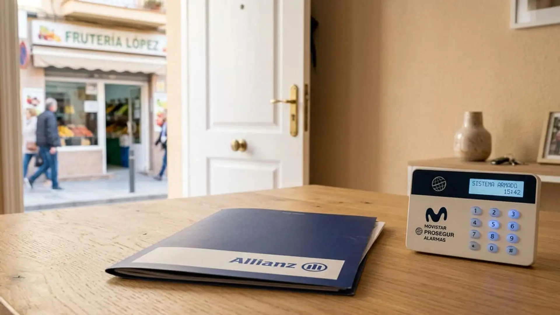Carpeta de Allianz junto a un teclado de alarma Movistar Prosegur sobre una mesa, con una frutería al fondo.