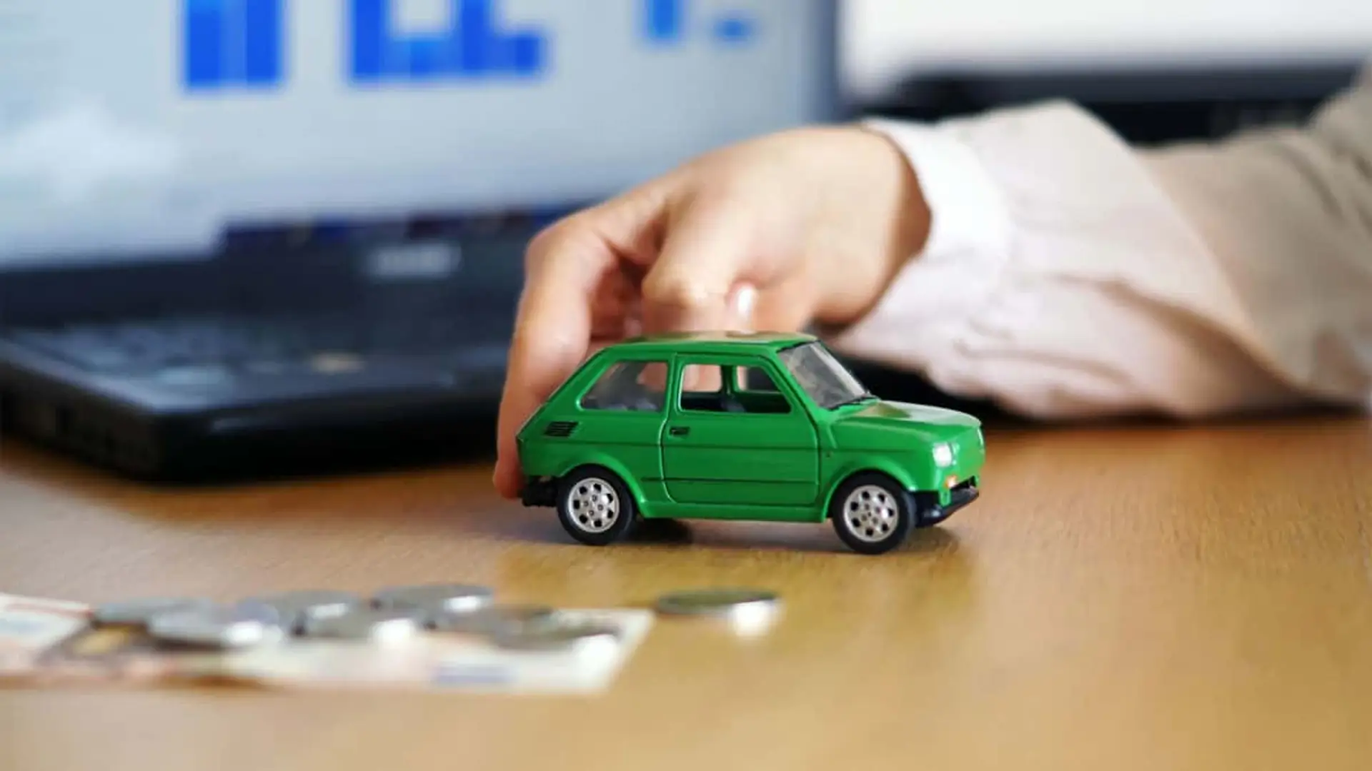 Persona calculando el precio de un seguro de coche con un coche de juguete, dinero y un portátil de fondo.