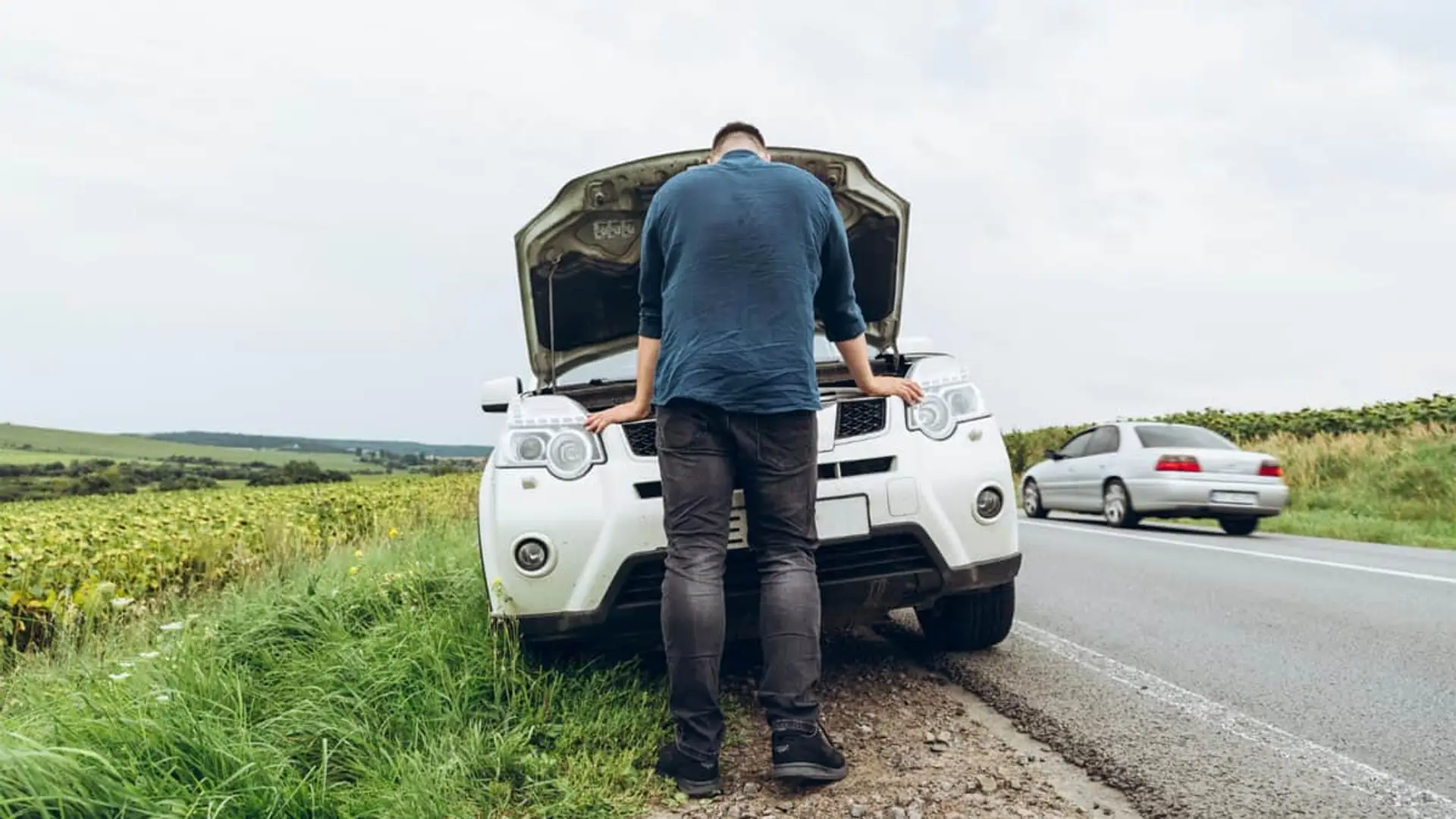 Conductor revisando motor de coche blanco averiado en el arcén de carretera con tráfico.