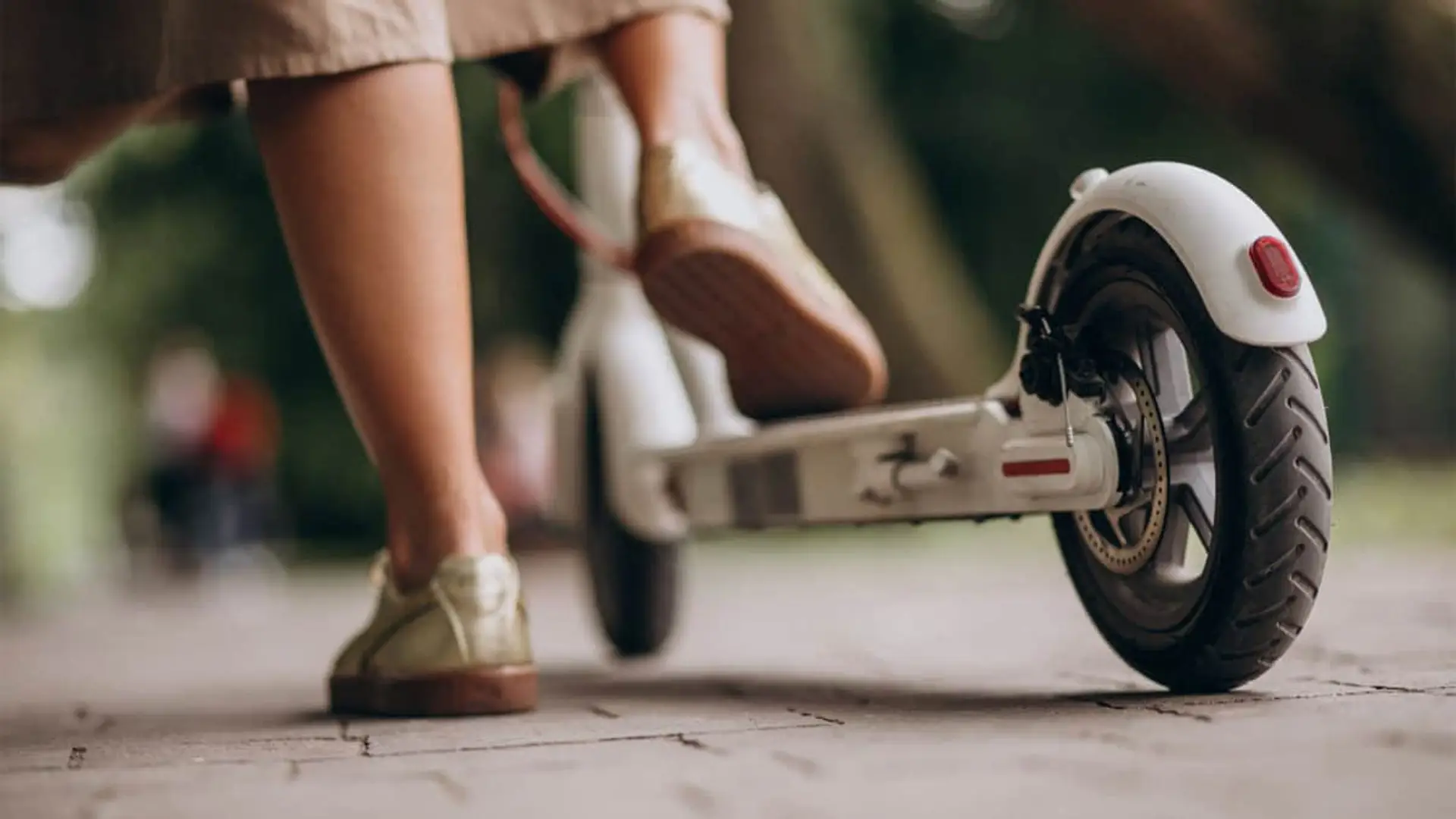 Ni seguro ni matrícula para patinetes eléctricos en enero: el lío legal de la DGT que cambia las normas en 2026 Primer plano de la rueda trasera y pies de una mujer conduciendo un patinete eléctrico por la calle.
