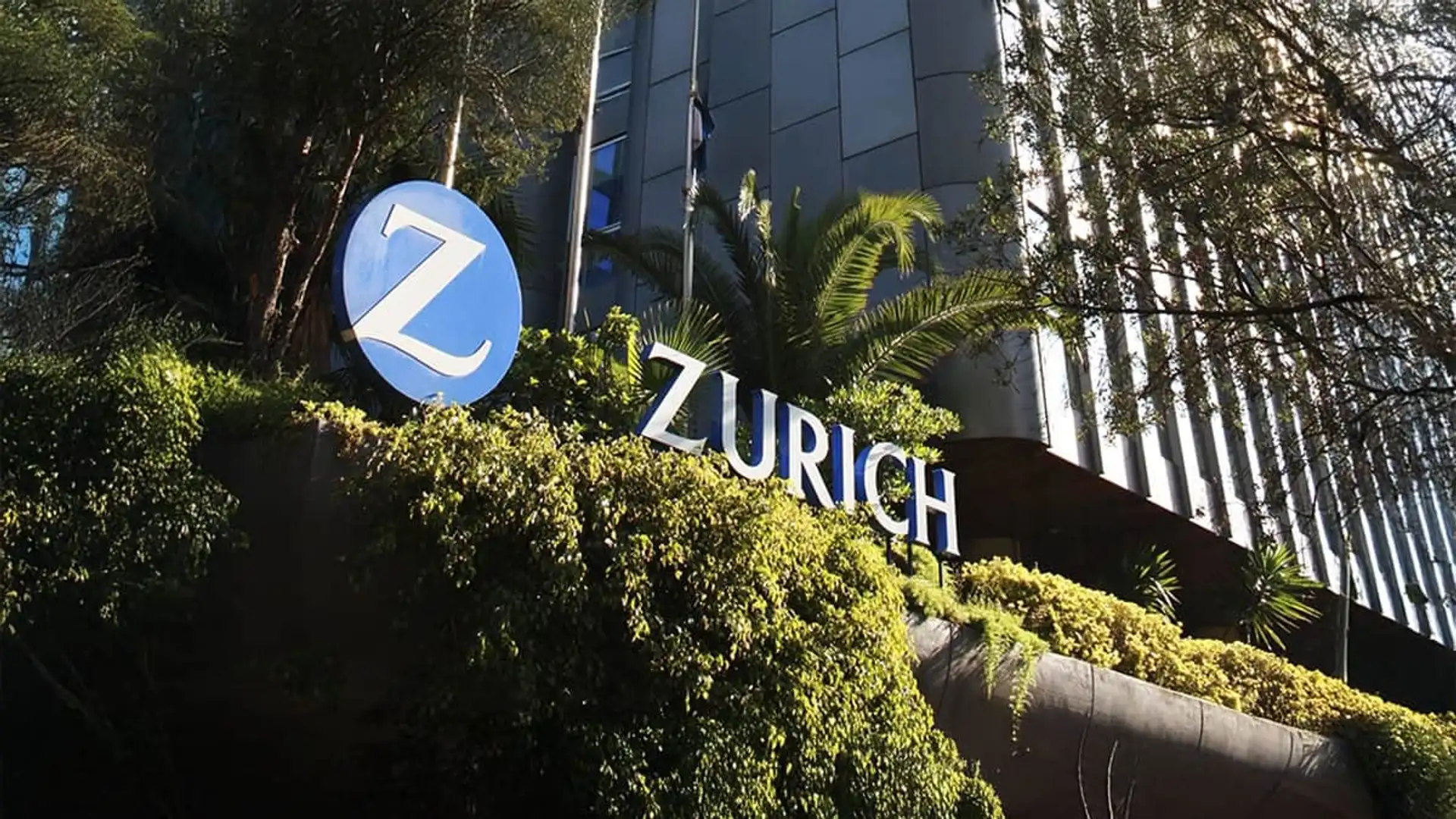 Logotipo de Zurich Seguros en la fachada de su sede corporativa rodeada de árboles y vegetación.