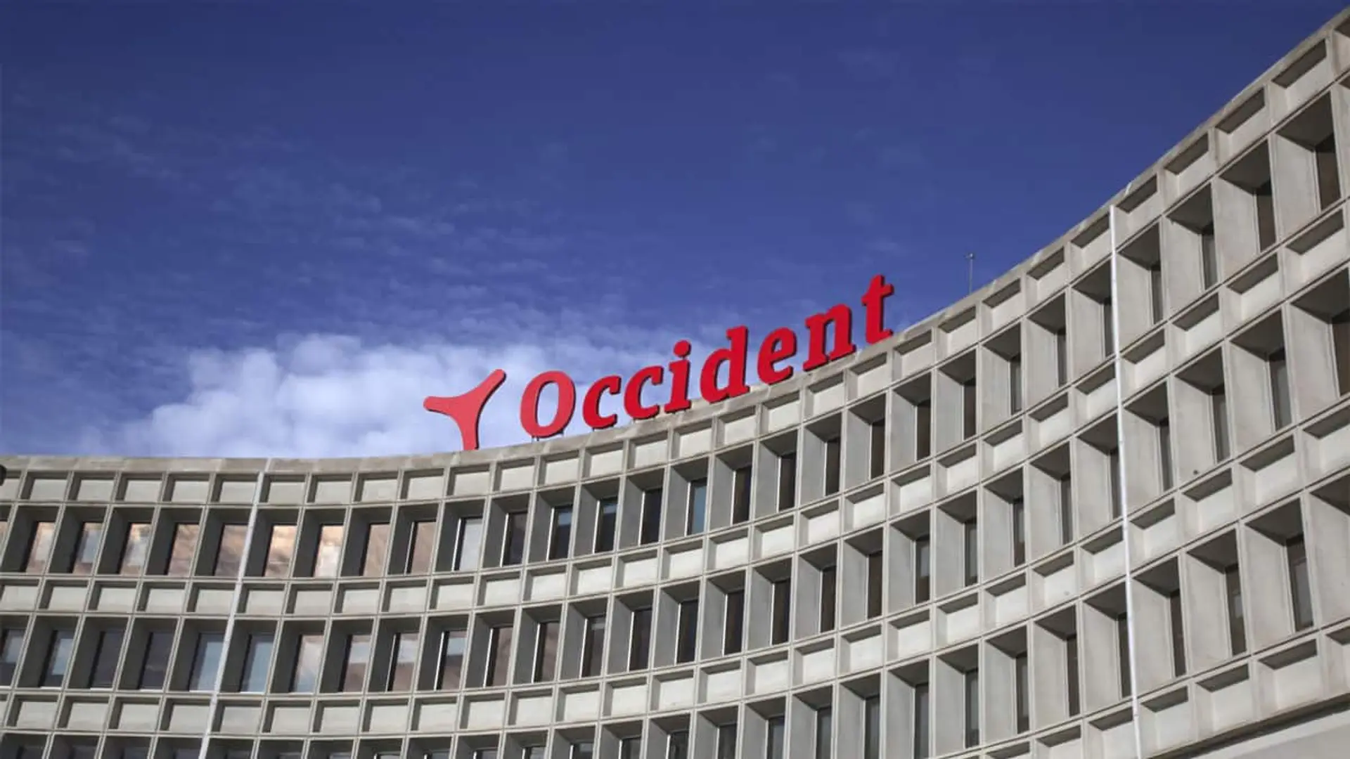 Catalana Occidente mira a decesos: quiere que la primera funeraria de España sea más grande y tiene 900 millones para ello Fachada del edificio principal de Occident Seguros con su logotipo rojo visible sobre fondo azul despejado.