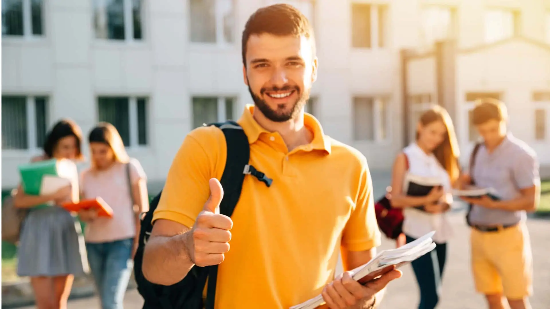 Joven estudiante sonriente en un campus universitario con camiseta amarilla y mochila, mostrando el pulgar hacia arriba y sosteniendo cuadernos.