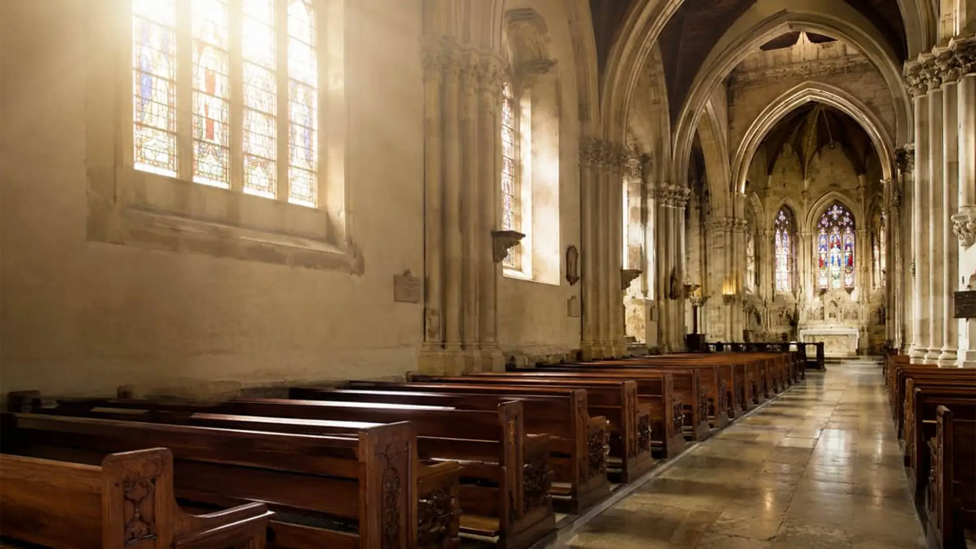 Interior de iglesia gótica con bancos de madera, vidrieras y luz natural sobre el altar