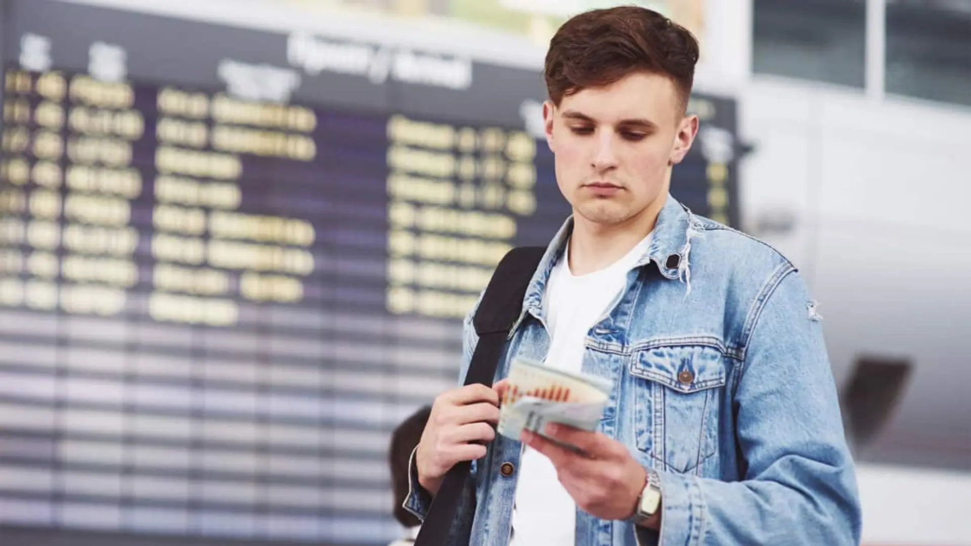 Joven viajero revisando billetes en un aeropuerto frente al panel de salidas