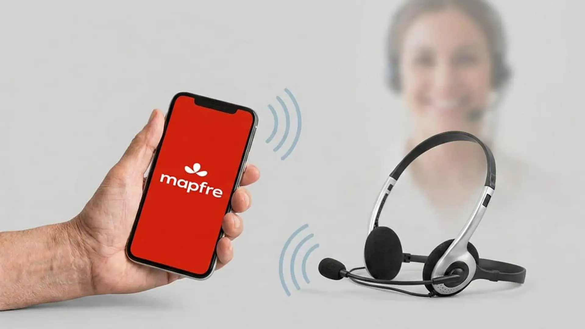 Mano sosteniendo un móvil con el logotipo de Mapfre junto a unos auriculares de atención al cliente y una operadora desenfocada al fondo