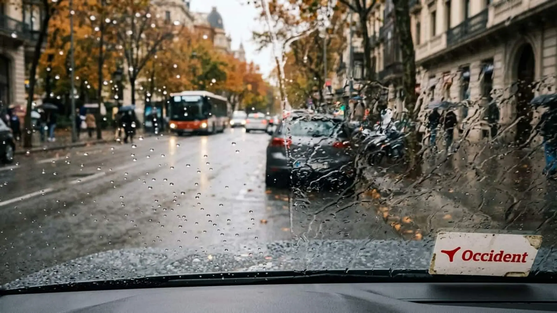 Occident regala un tratamiento antilluvia si tienes garantía de lunas: cómo pedirlo hasta el 30 de noviembre Vista desde el interior de un coche bajo la lluvia con tráfico urbano y el logotipo de Occident en el parabrisas