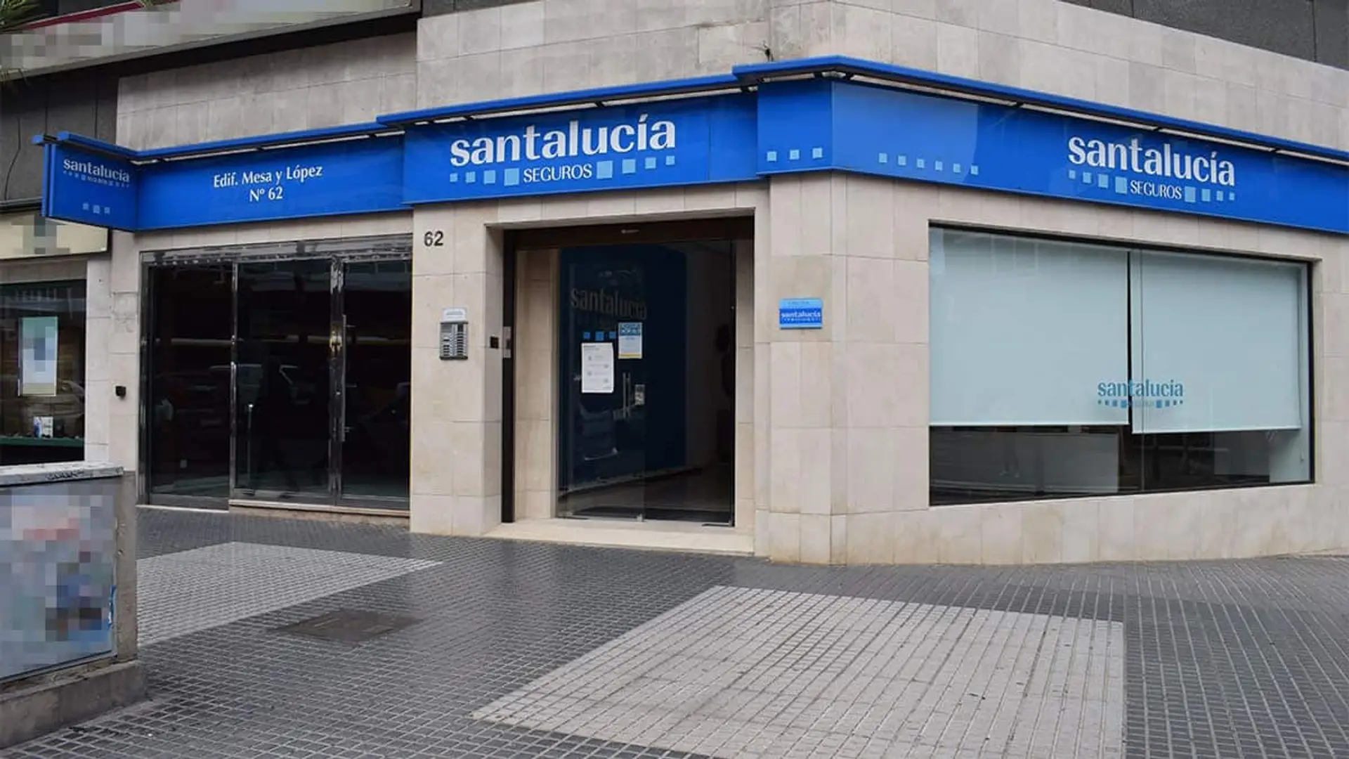 Santalucía lanza tres nuevos seguros de decesos flexibles y personalizados Fachada de una oficina de Santalucía Seguros en una esquina de calle con rótulos azules.