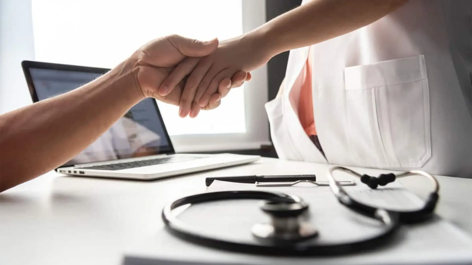 Paciente y profesional sanitario dándose la mano en una consulta con un fonendoscopio sobre la mesa