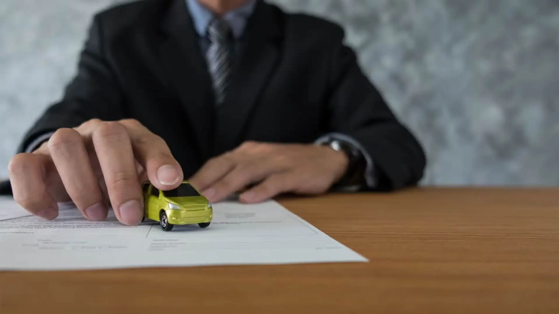 ¿Te han subido el precio del seguro de coche? Estos son los más baratos en noviembre de 2025 Persona formalizando un contrato de seguro de coche. Su mano coloca un vehículo en miniatura sobre la póliza en el escritorio.