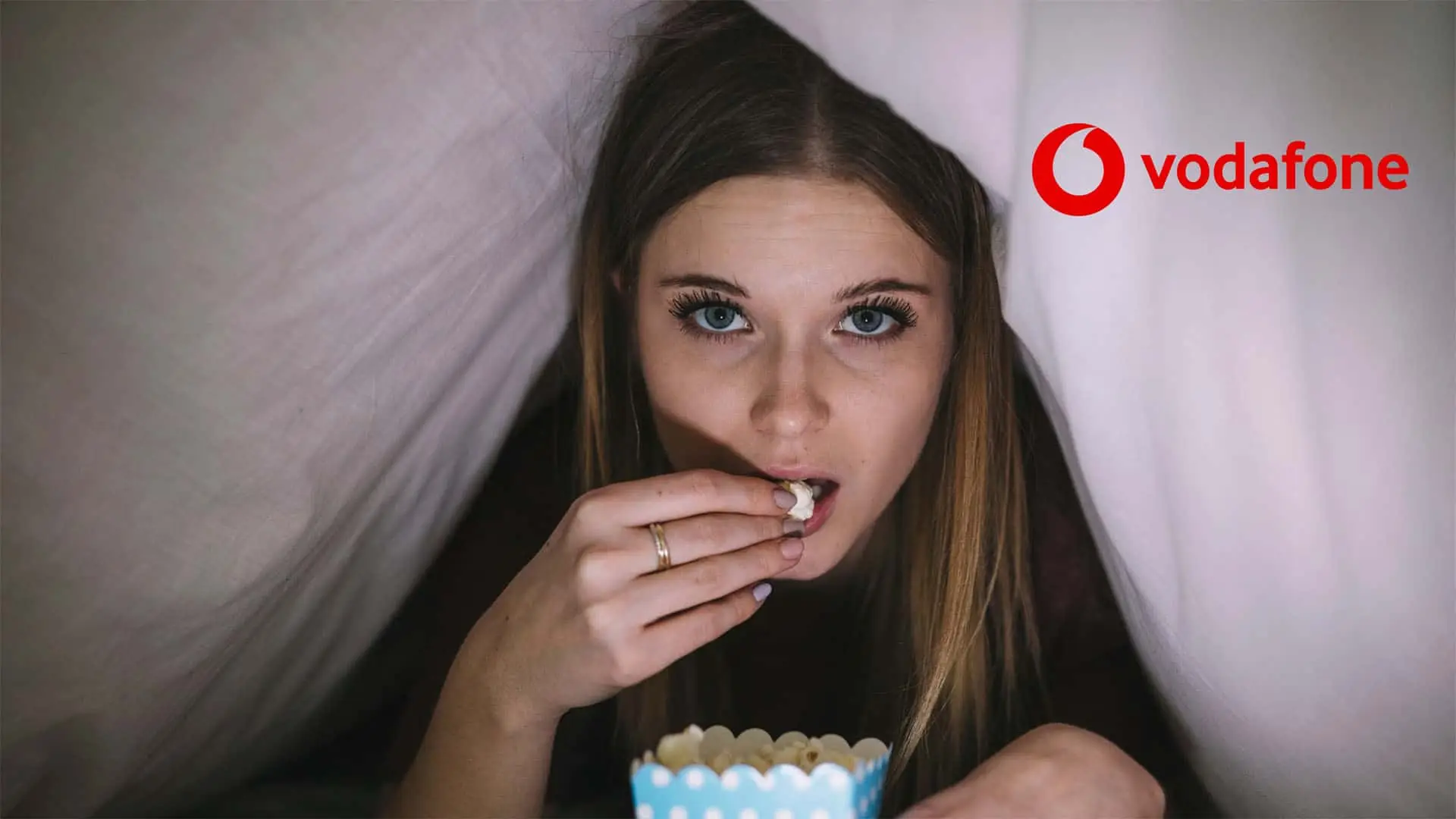 Joven viendo una película de terror en la cama. Se esconde bajo las sábanas mientras come palomitas.