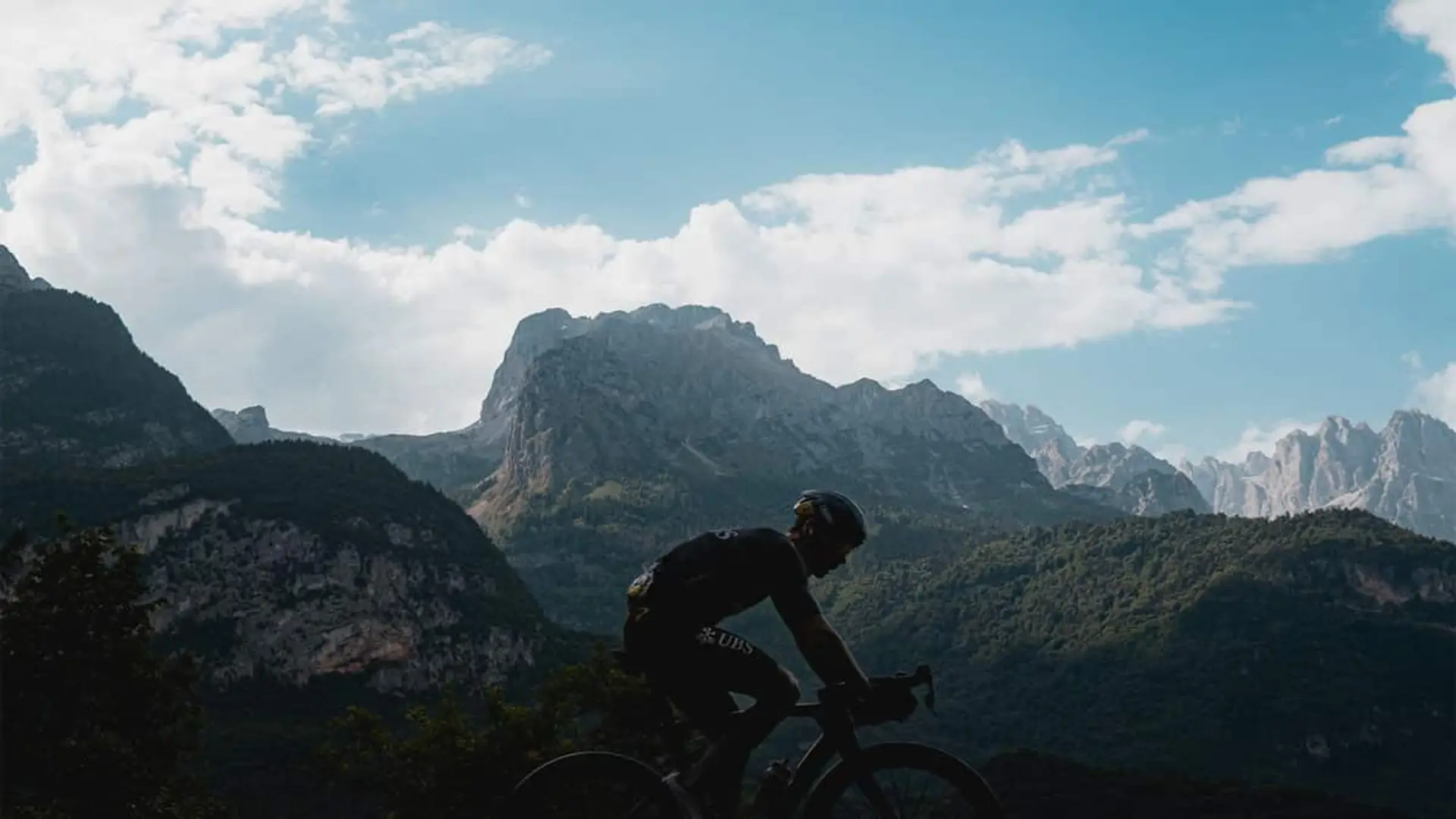 Ciclista en silueta pedaleando en el Giro de Italia por una carretera de montaña con un macizo montañoso al fondo