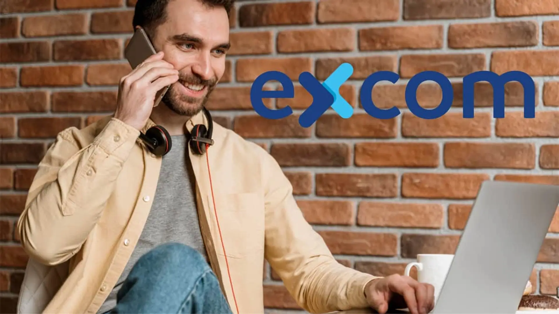 MasOrange y Vodafone pujan por Excom: por esto les interesa tanto esta operadora local Hombre sonriente usando su portátil y hablando por móvil, con el logo de la operadora Excom.