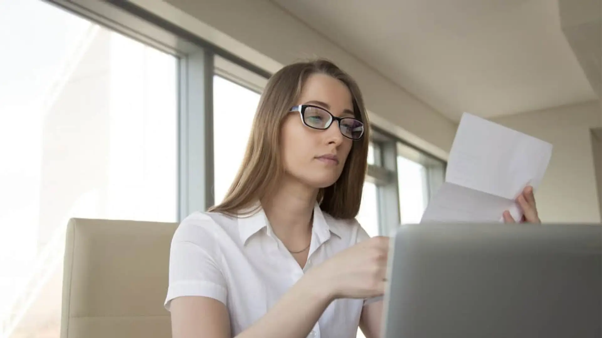 Orange subirá los precios en 2026: prepara aumentos de hasta 72€ anuales en sus tarifas de fibra, móvil y TV Mujer revisando documento o factura en papel frente al ordenador portátil en la oficina.