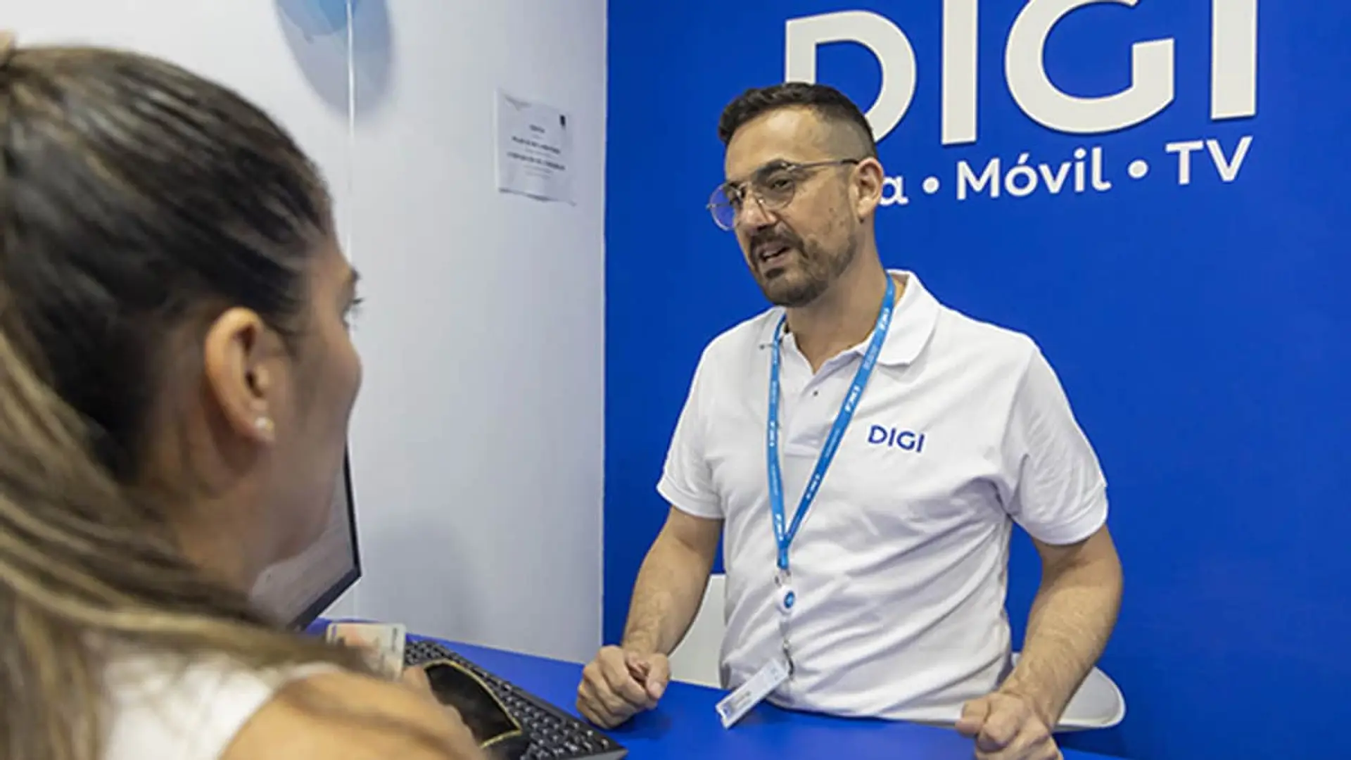 Por qué DIGI sube su penalización a 100 euros: la estrategia para frenar a los cazadores de ofertas Asesor de DIGI atendiendo a una clienta en una tienda de fibra, móvil y TV con el logotipo de la marca al fondo.