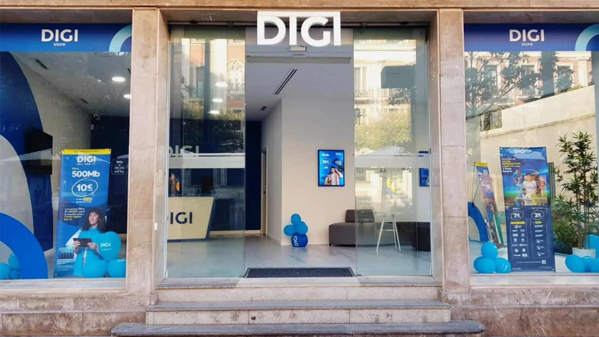 DIGI saldrá a Bolsa en España para seguir creciendo: el Santander y otros bancos le ayudarán Fachada exterior de una tienda DIGI con carteles publicitarios y globos corporativos azules.