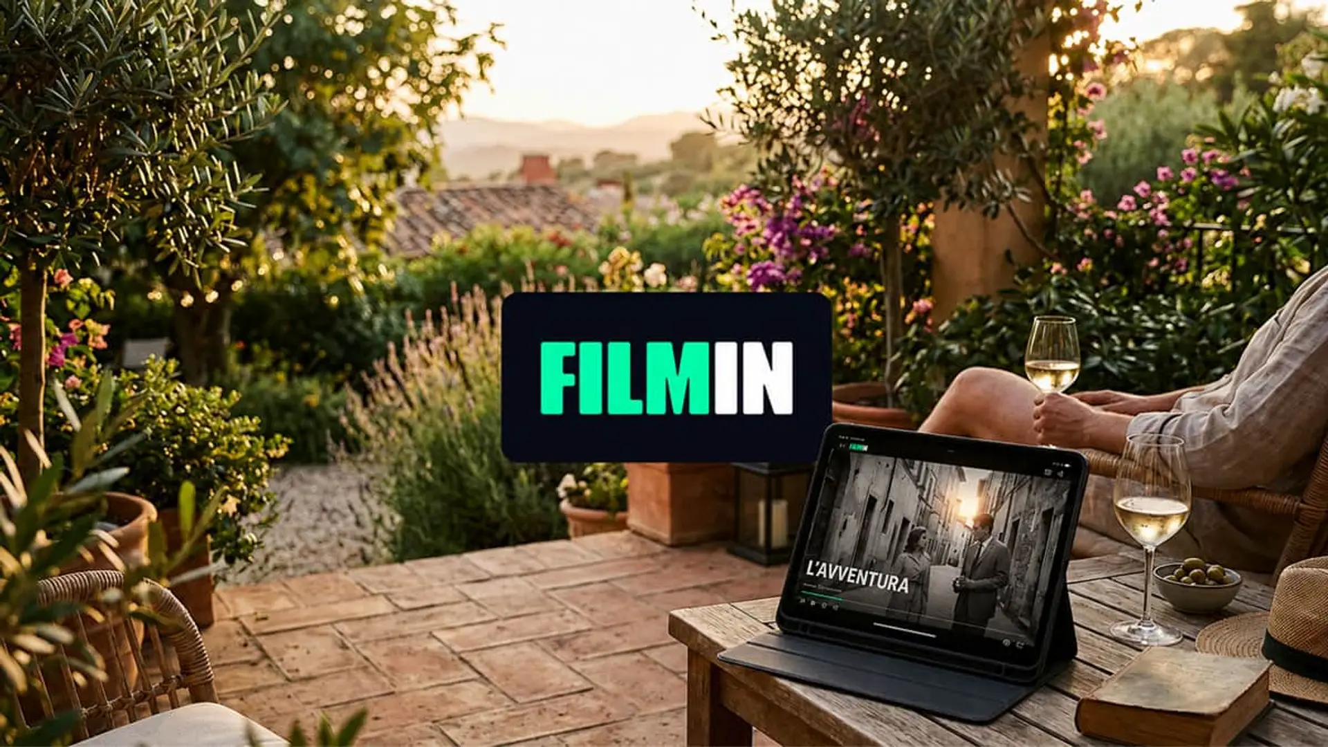 Filmin se suma a las rebajas de verano en streaming: baja sus precios hasta el 3 de agosto Filmin en una terraza mediterránea, con una película en una tablet, copas de vino y jardín al atardecer.