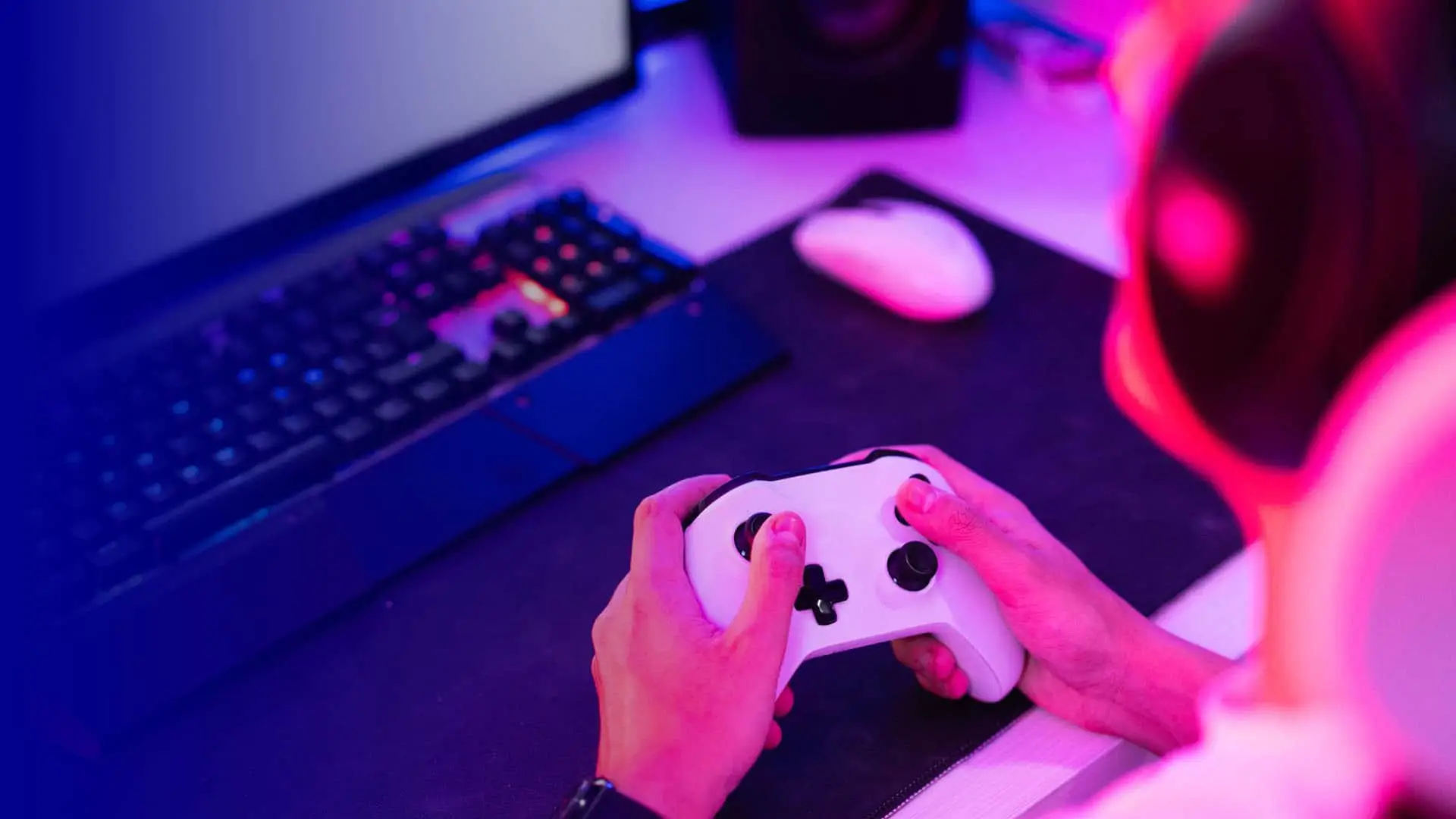 Persona jugando videojuegos con un mando blanco frente a un PC gaming con teclado mecánico y luces LED.