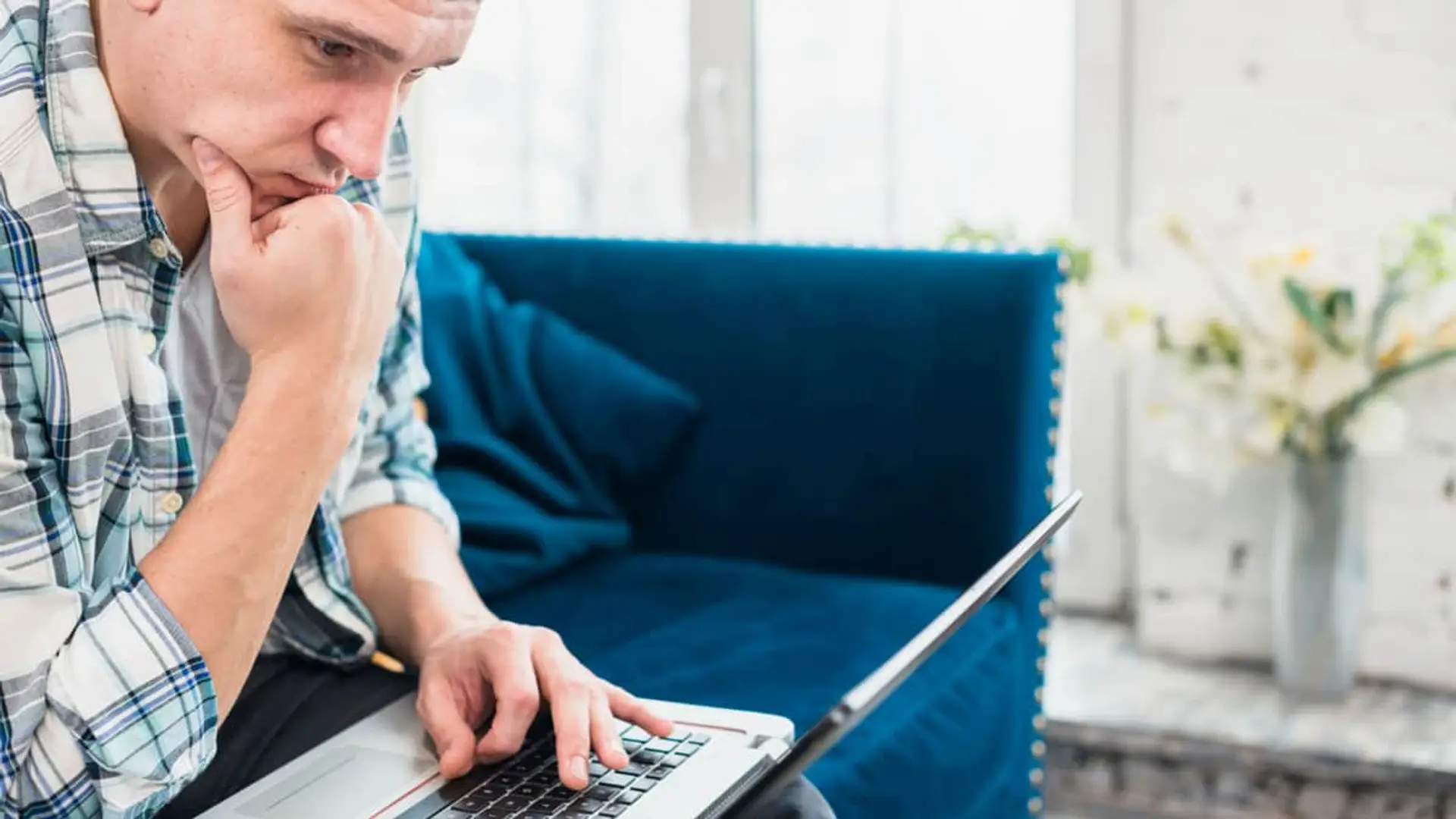 Hombre concentrado usando un ordenador portátil en el sofá de casa para gestiones online o teletrabajo.