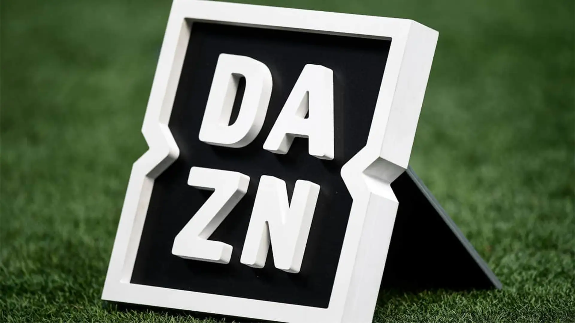 DAZN hunde sus precios en España: nuevas ofertas para ver el fútbol, el baloncesto y la F1 Logo corpóreo de la plataforma de streaming DAZN sobre el césped de un campo de fútbol.