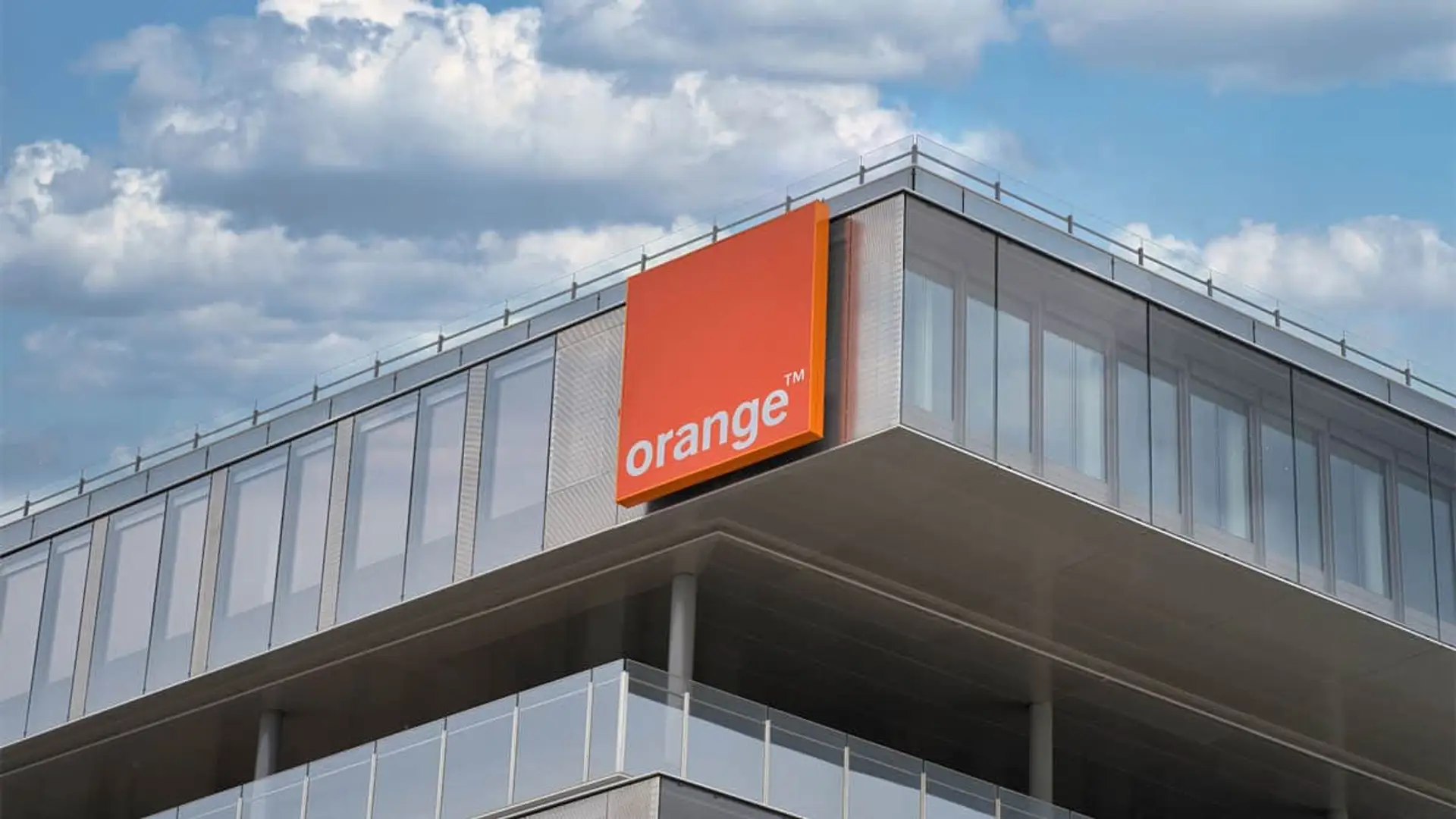 Francia ya posee el 100% del operador líder en España: así ha tomado el control total de MasOrange Edificio corporativo con letrero naranja de Orange sobre fachada de vidrio y metal, cielo azul con nubes.