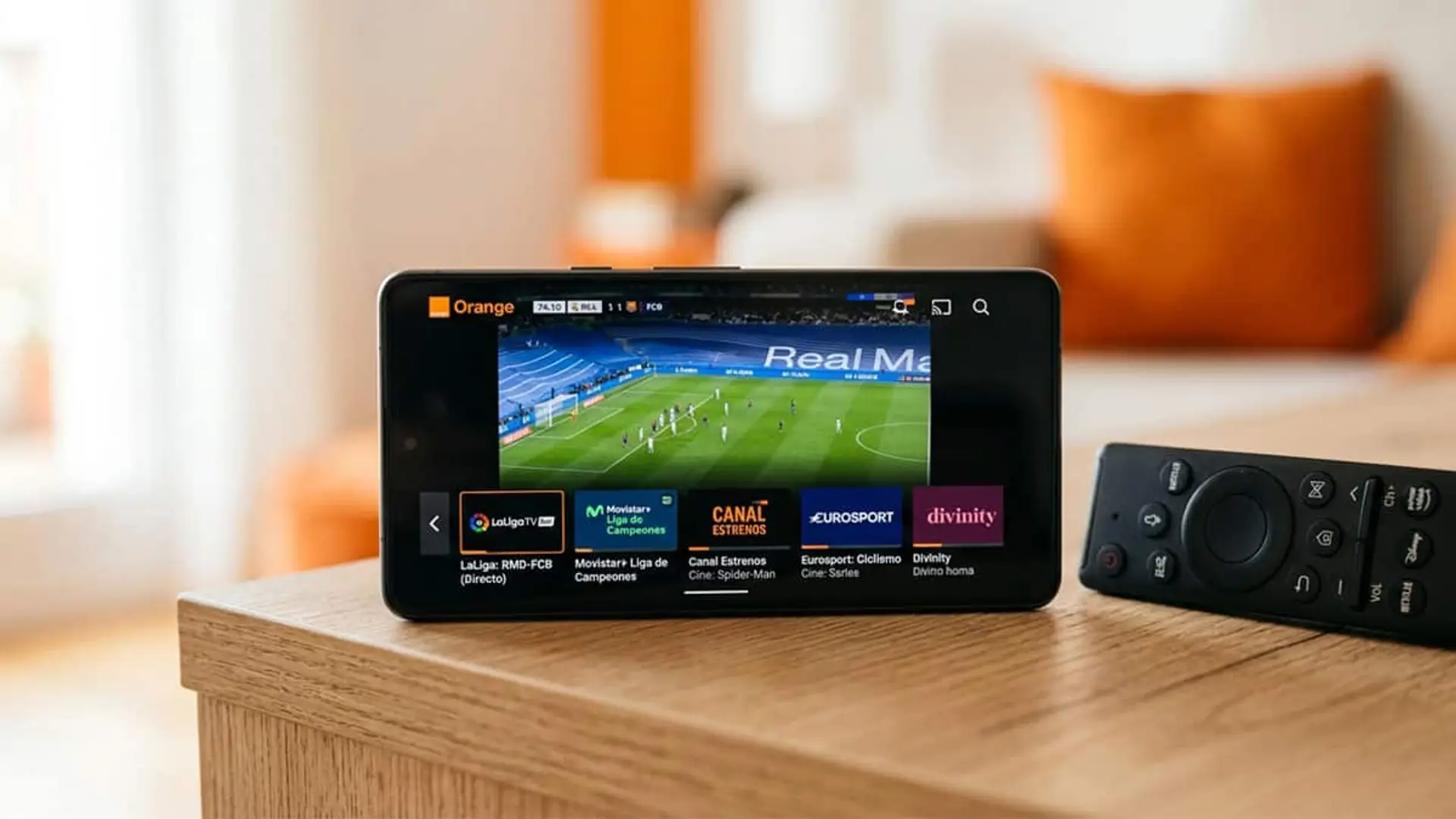Móvil con Orange TV mostrando un partido de fútbol y accesos a varios canales junto a un mando a distancia