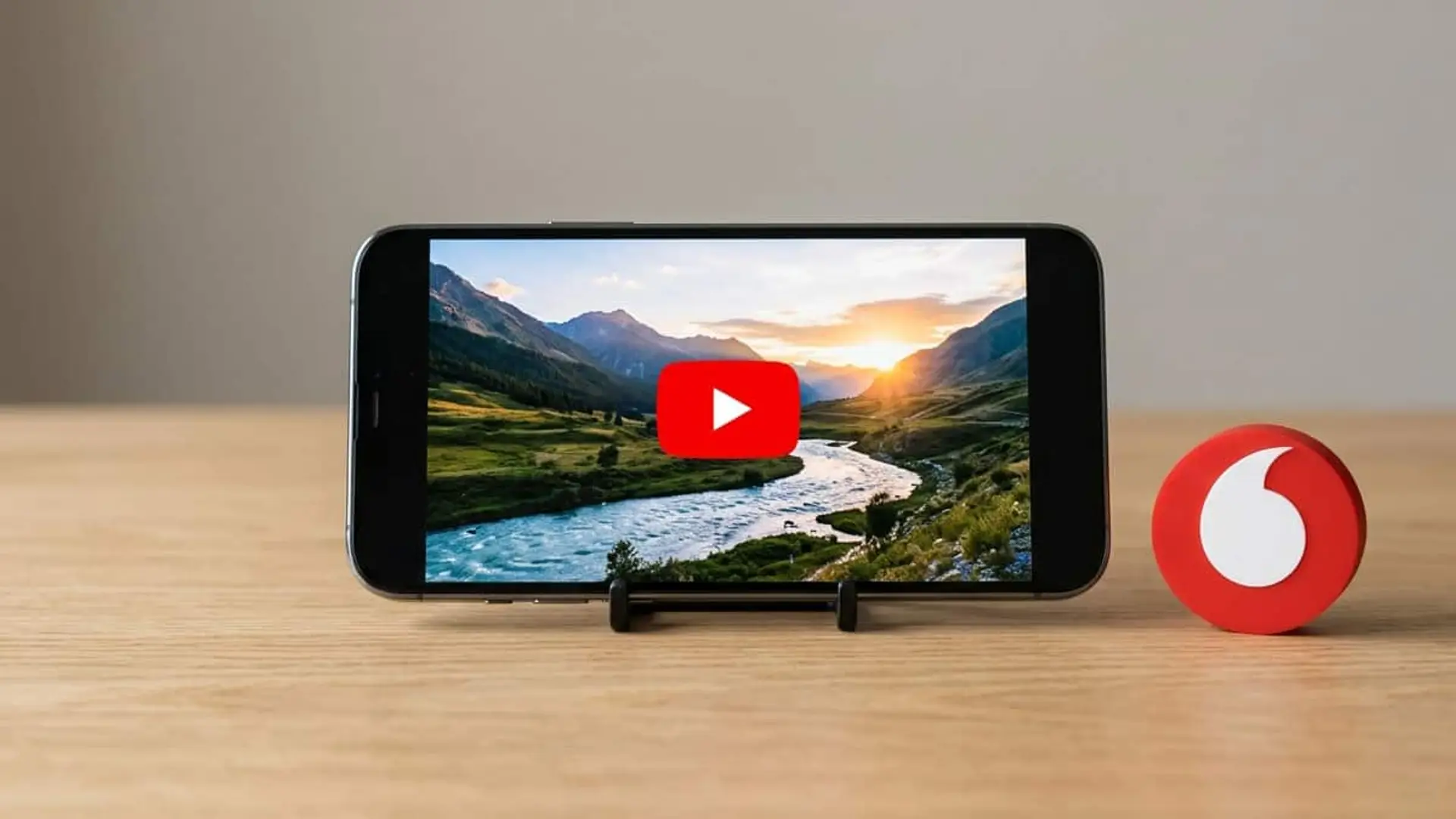 YouTube Premium Lite o Premium con Vodafone: 3 euros menos al mes y qué pierdes Teléfono móvil en horizontal mostrando el logotipo de YouTube sobre un vídeo de un paisaje de montañas y un río, junto al logotipo rojo de Vodafone sobre una mesa de madera.