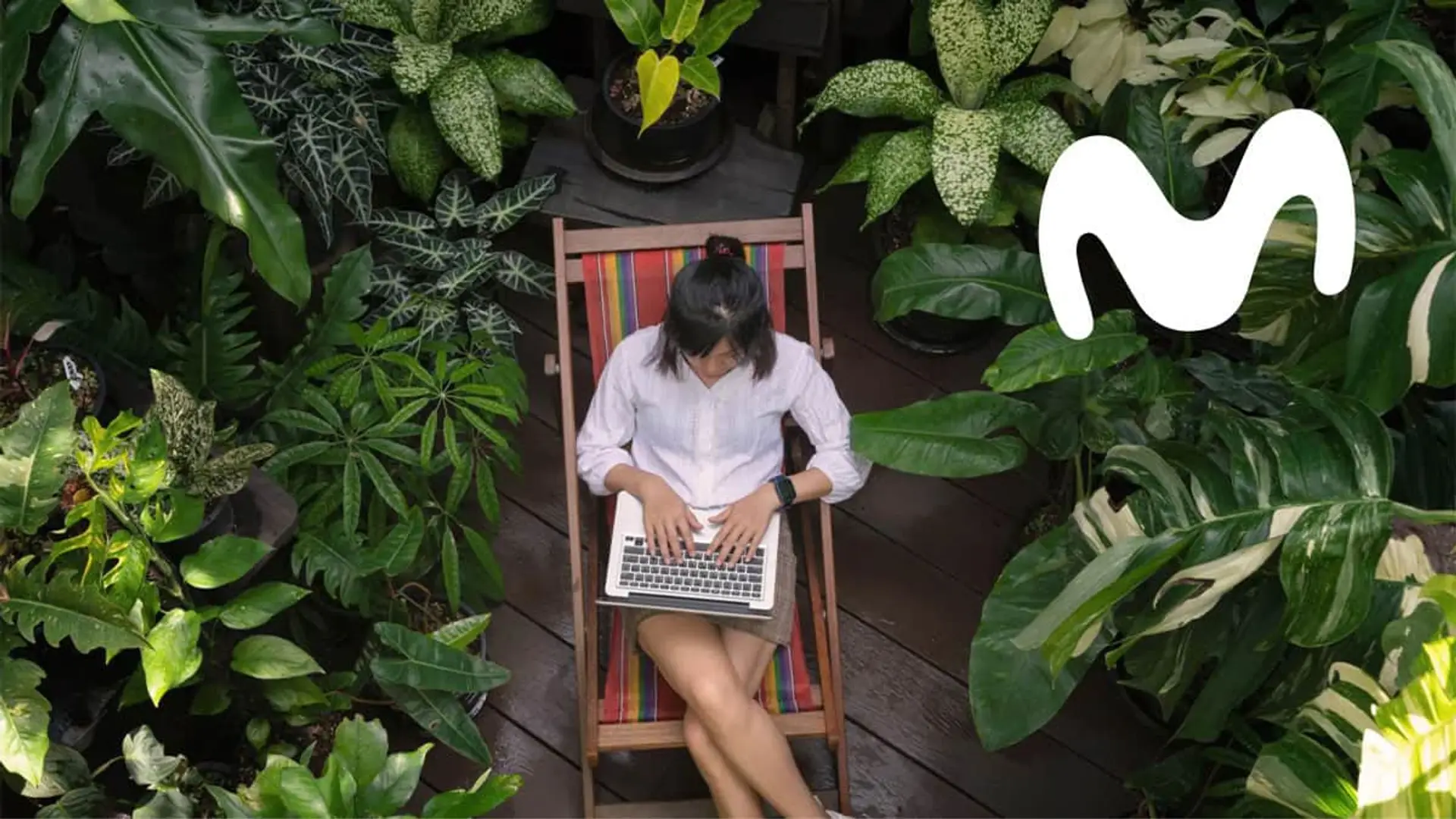 Mujer usando un portátil en un jardín junto al logo de Movistar para representar el teletrabajo y la conectividad.