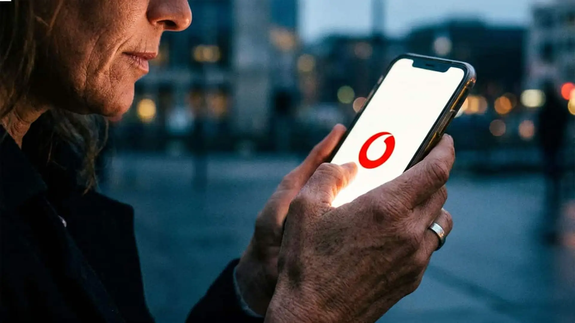 Vodafone confirma su subida de precios en 2026: se desliga del IPC y sigue los pasos de Movistar y Orange Primer plano de una mujer usando su móvil al aire libre por la noche con el logotipo de Vodafone en la pantalla.