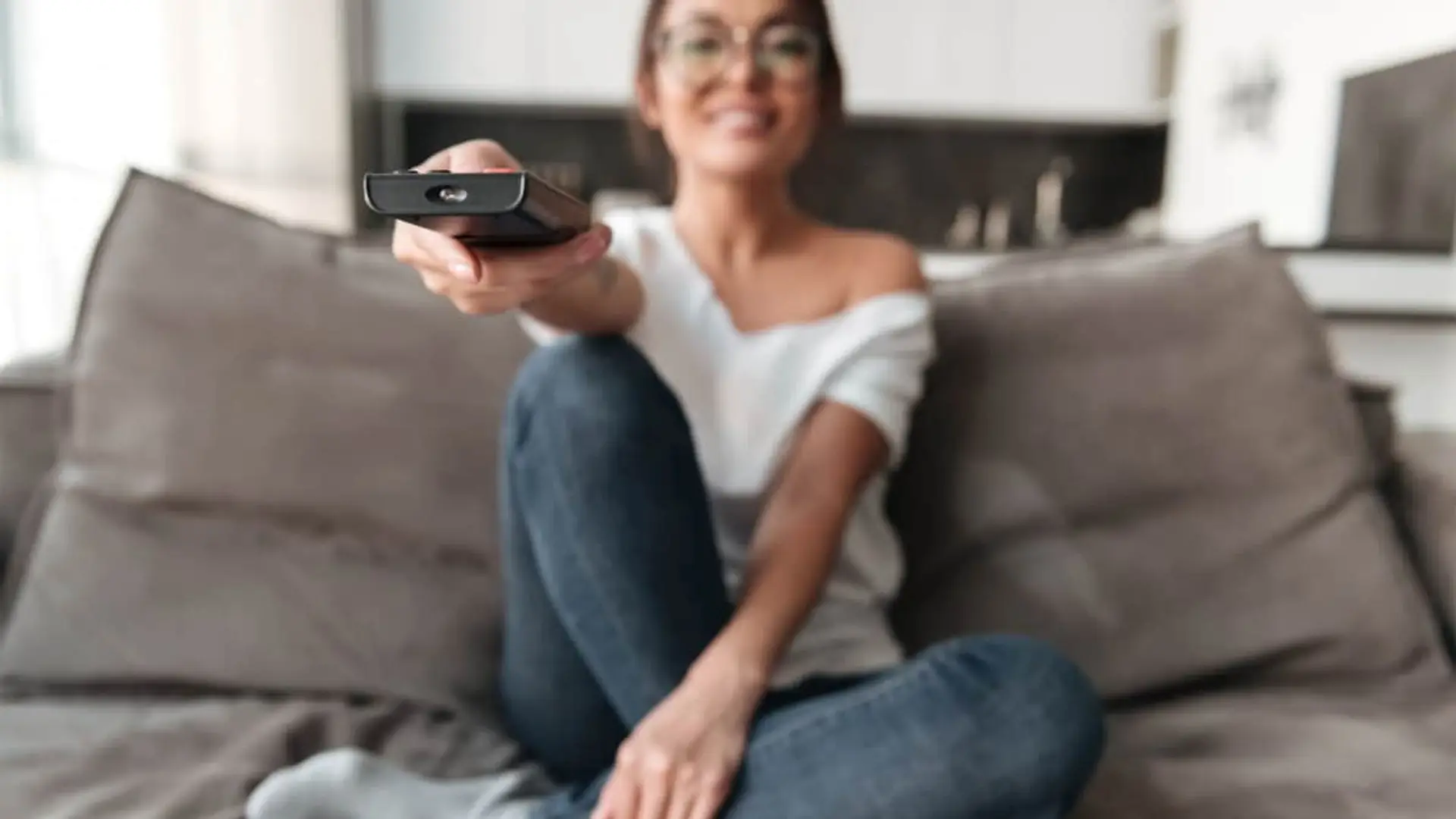 Estrenos del 14 al 16 de noviembre en Netflix, Prime Video, HBO Max y Disney+: qué ver este fin de semana Primer plano de un mando a distancia. Al fondo, una mujer sonriente con gafas sentada en el sofá para ver la tele.